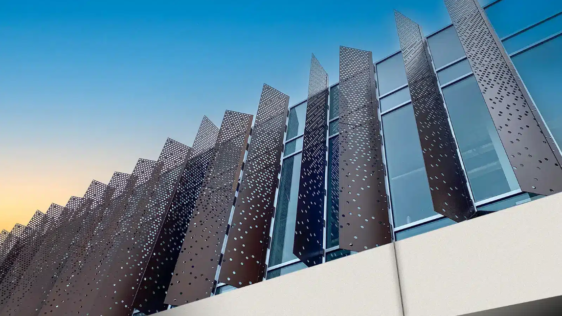 Modern building exterior with perforated metal panels and glass windows, viewed against a blue sky during sunset.