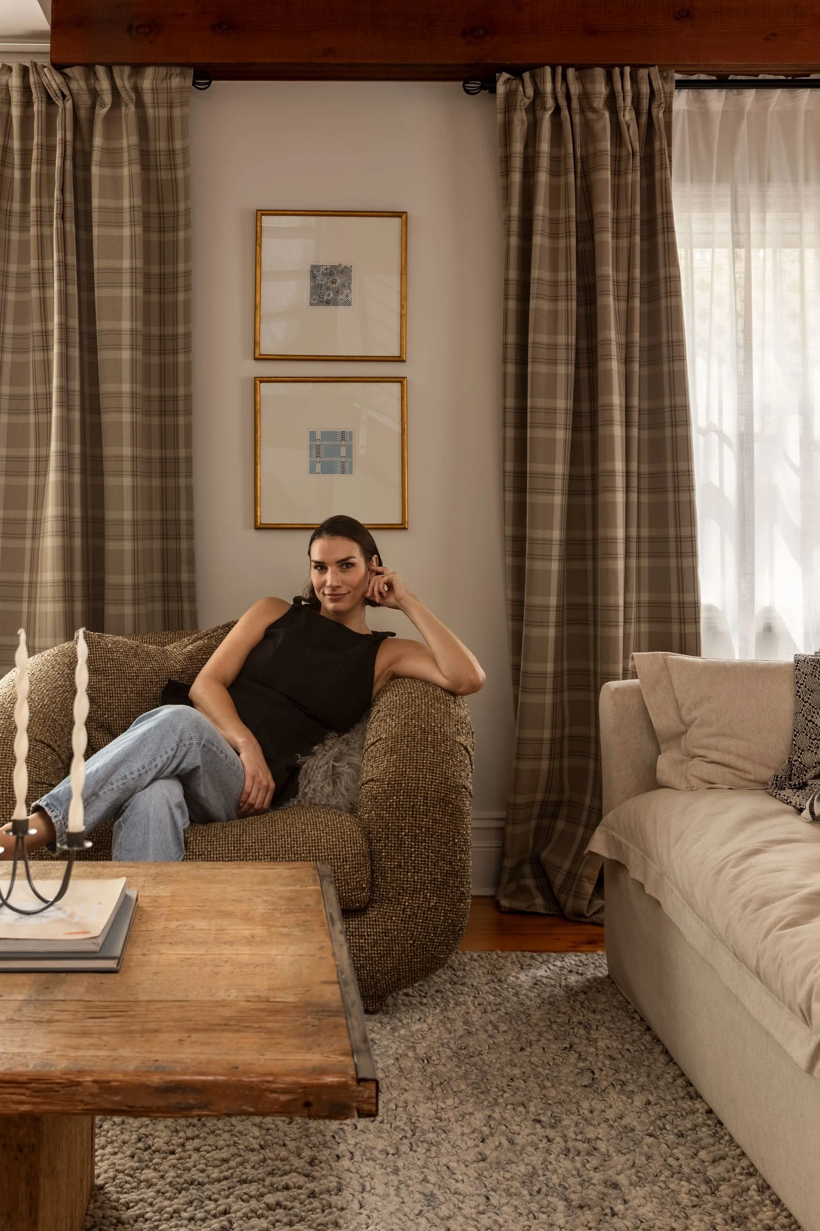 A woman sitting on a brown couch in a cozy living room, with beige curtains and framed artwork on the wall behind her, and a wooden coffee table in front of her.