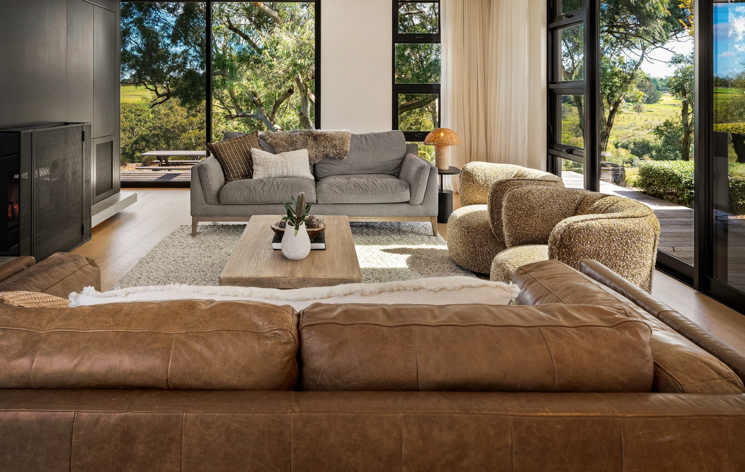 Living room with large windows showing lush green trees and landscape outside. Features a gray sofa with cushions, a wooden coffee table with a white vase and plant, textured chairs, a multi-toned rug, side table with a lamp, and beige curtains.