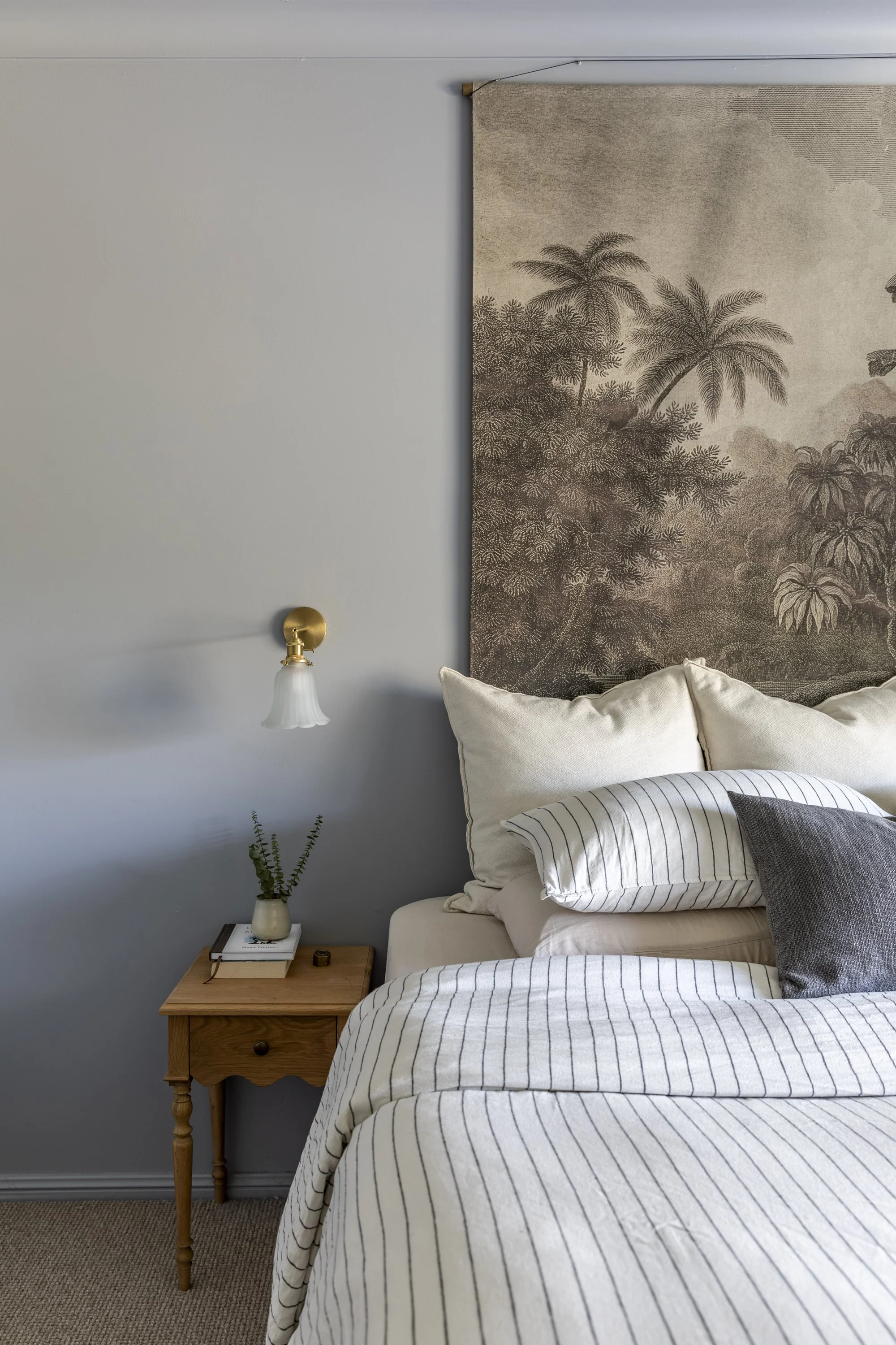 Bedroom with a bed, white and gray striped bedding, pillows, a wooden nightstand with a small plant, books, and a lamp, and a wall hanging with tropical plants.