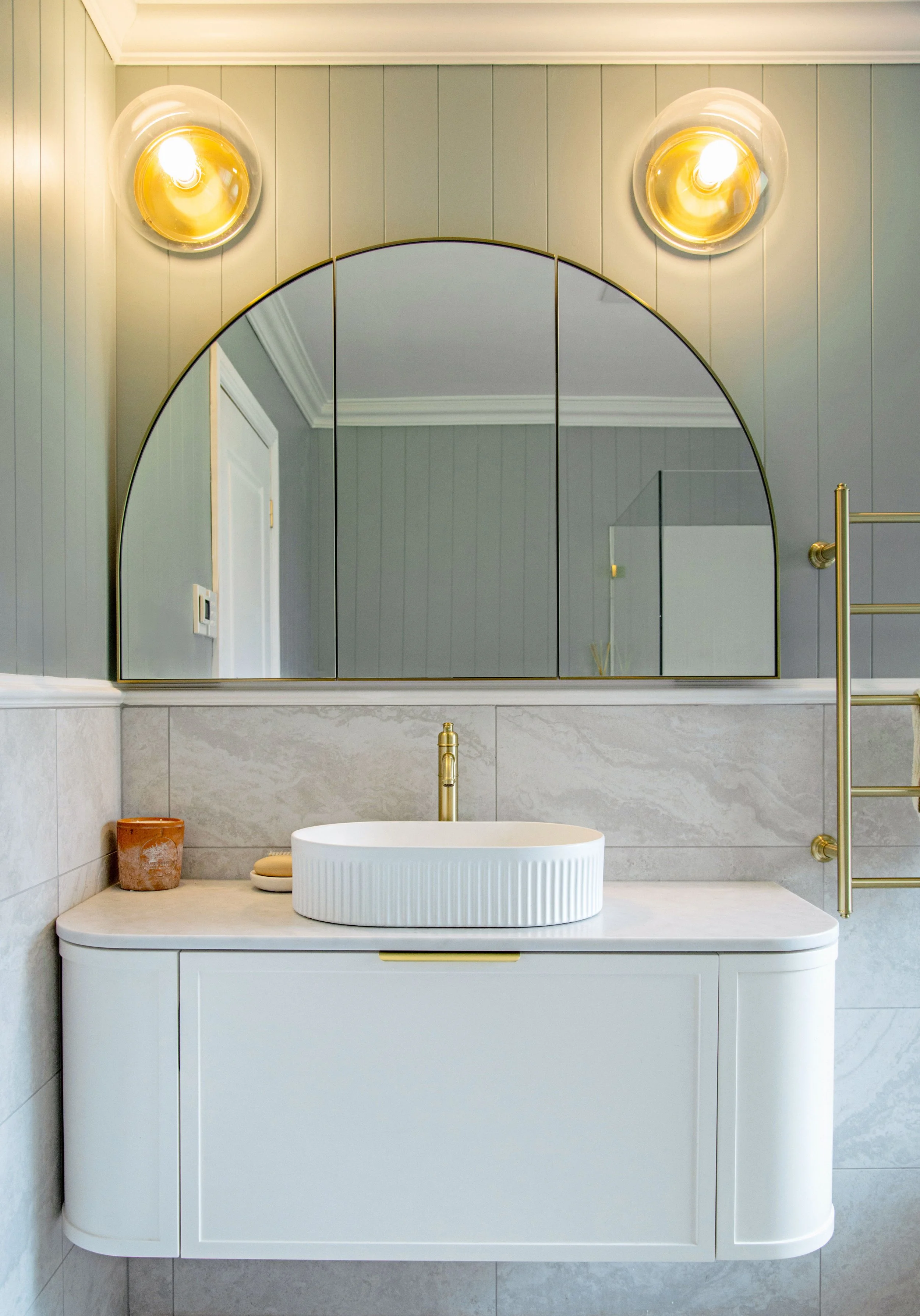 Bathroom with a large arched mirror above a white vanity with a vessel sink, gold fixtures, and minimalist decor including a small clay pot, a soap dish, and towel rack on the right.