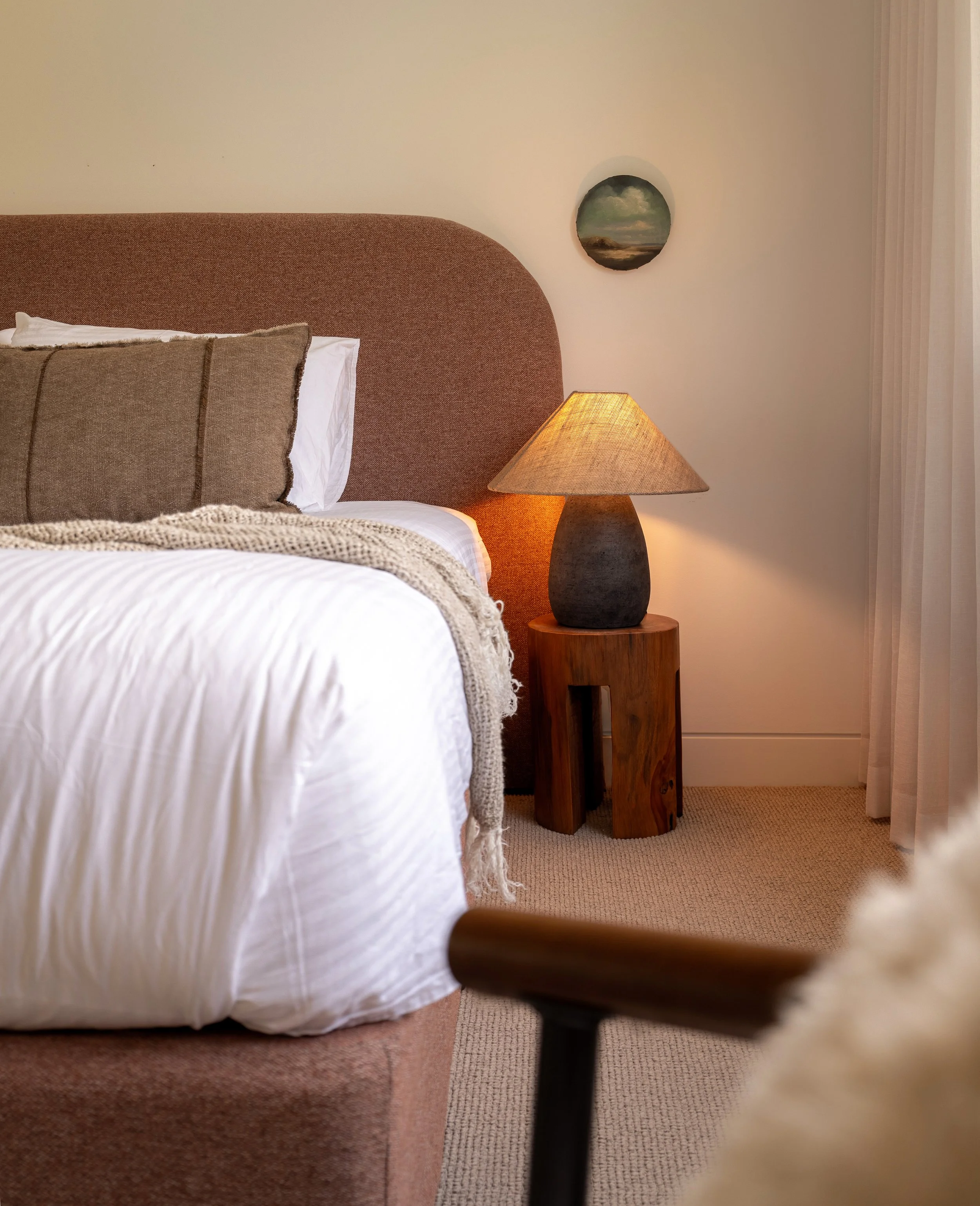 A bedroom with a bed that has a white duvet and beige pillows, a wooden nightstand with a textured brown lamp, a wall with a small landscape painting, and a curtain on the right side.