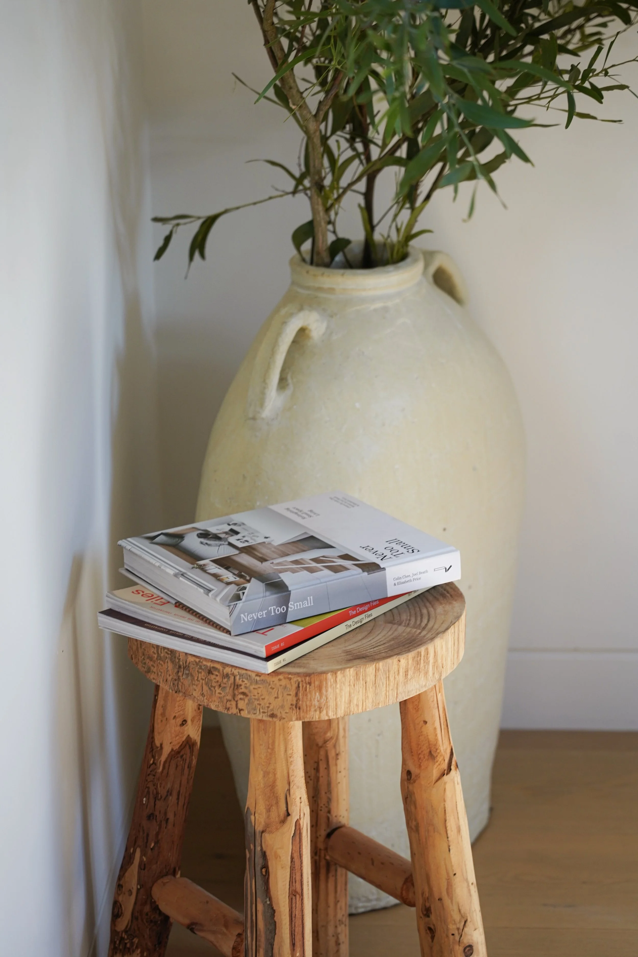 A rustic wooden side table with a round top, holding a stack of books and magazines. Behind the table, a large cream-colored ceramic vase with handles contains green leafy branches, set against a plain light-colored wall.