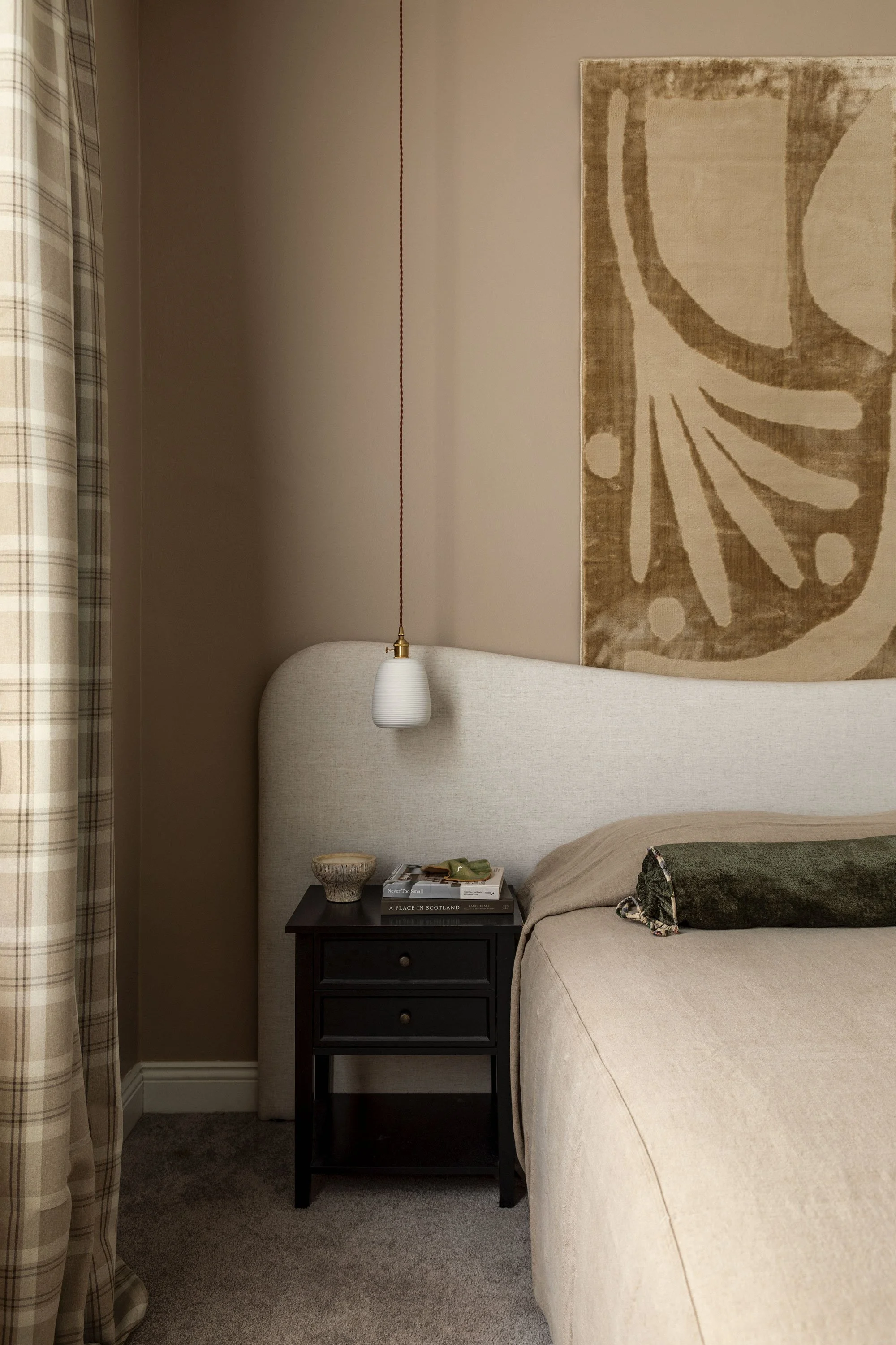 A bedroom corner with a beige upholstered bed, a dark wooden nightstand with books and a bowl, a white pendant light hanging from the ceiling, a large abstract art piece, and plaid curtains on the left side of the image.