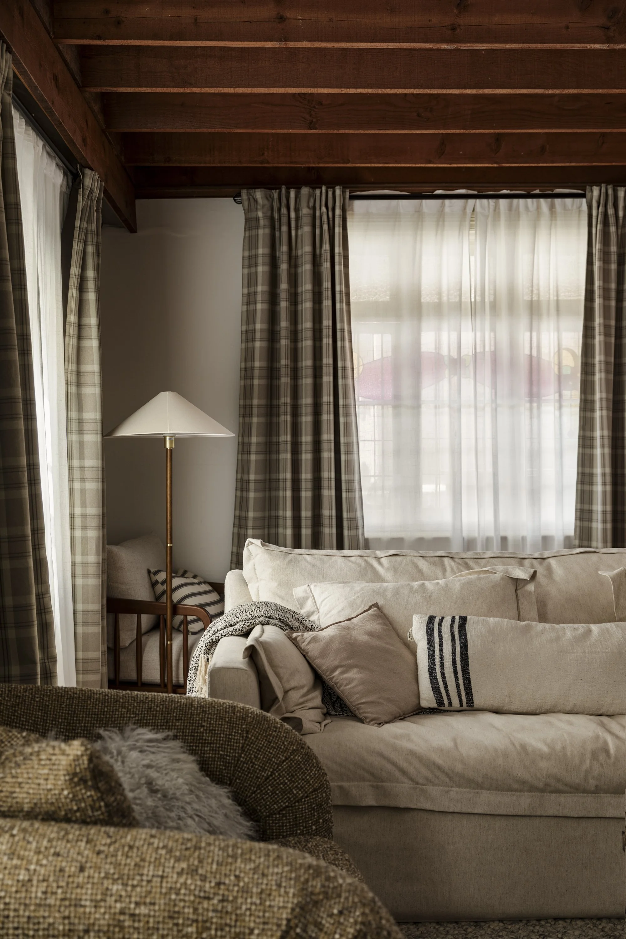 Cozy living room with beige sofa, plaid and striped pillows, wooden ceiling, large window with sheer curtains, and a standing lamp.
