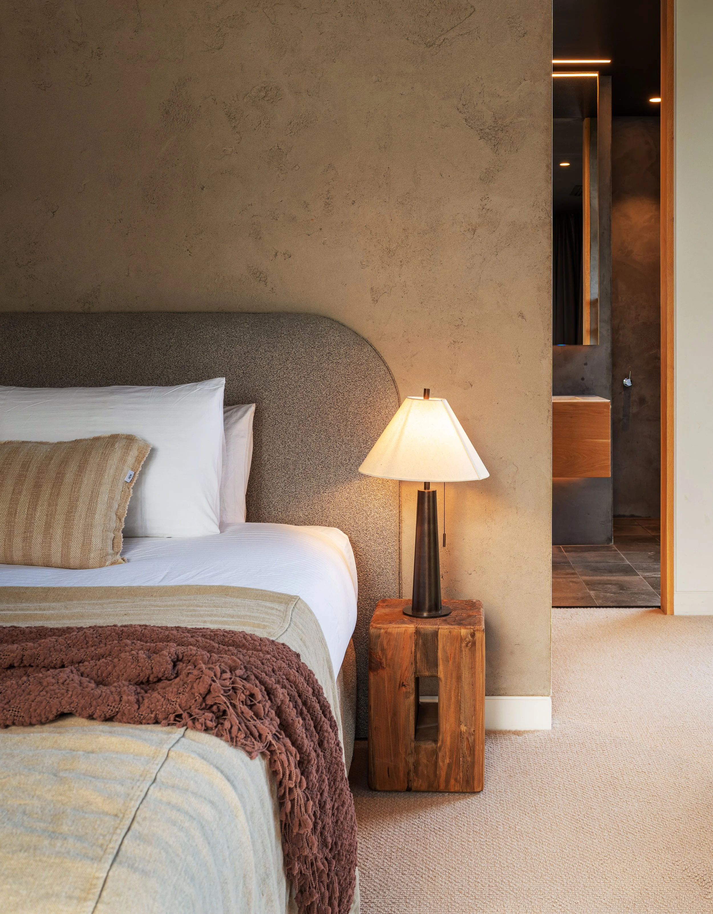 A cozy bedroom with a bed featuring white linens and a beige throw blanket, a textured concrete wall, a wooden nightstand with a black lamp, and a partial view of an open bathroom with dark gray walls and a mirror.
