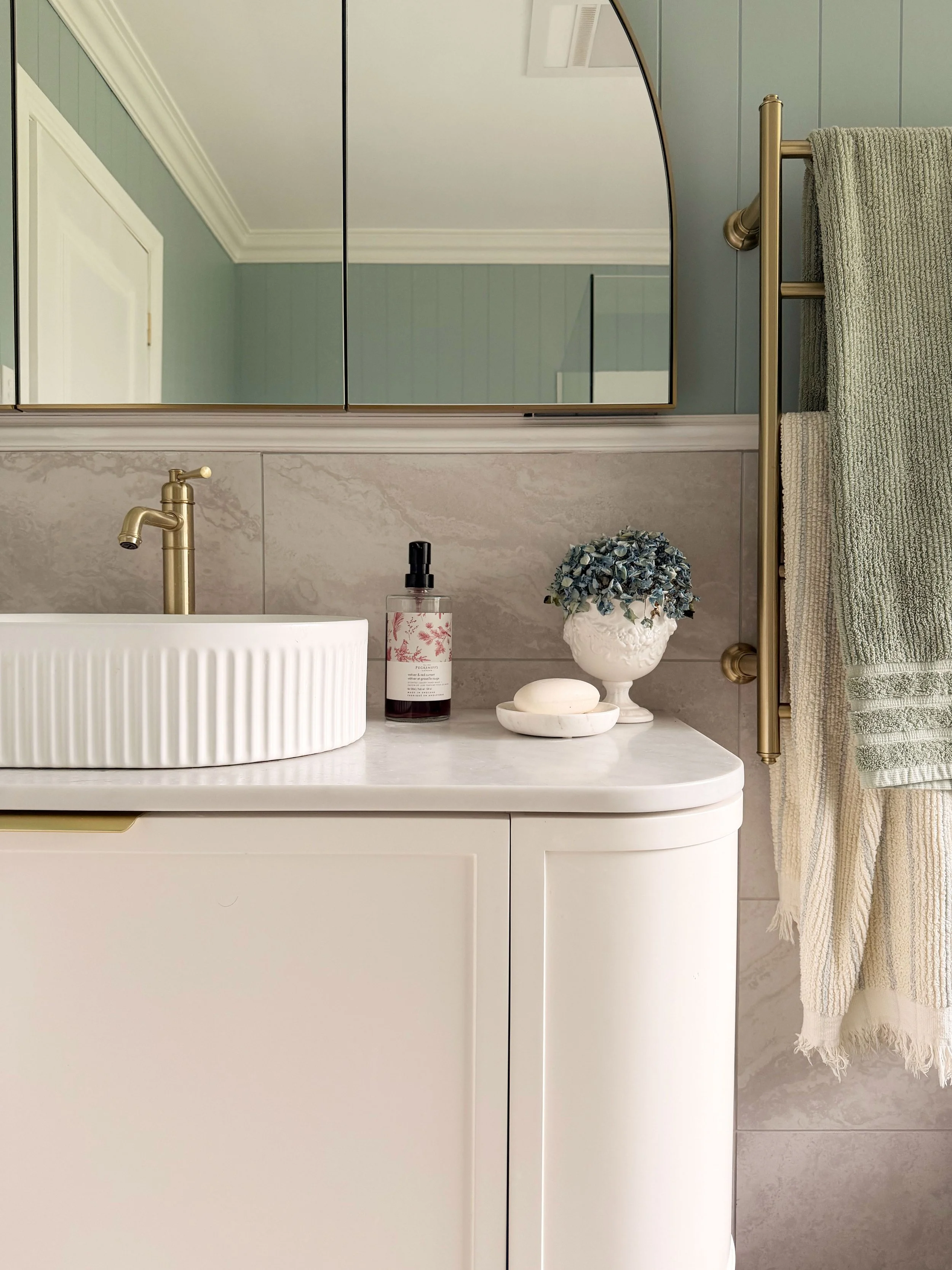 Bathroom with a white vanity, a round vessel sink with gold faucet, a soap dispenser, a white soap dish with soap, a vase with blue hydrangea, and beige towels hanging on a gold towel rack, mirror reflecting the ceiling and door.