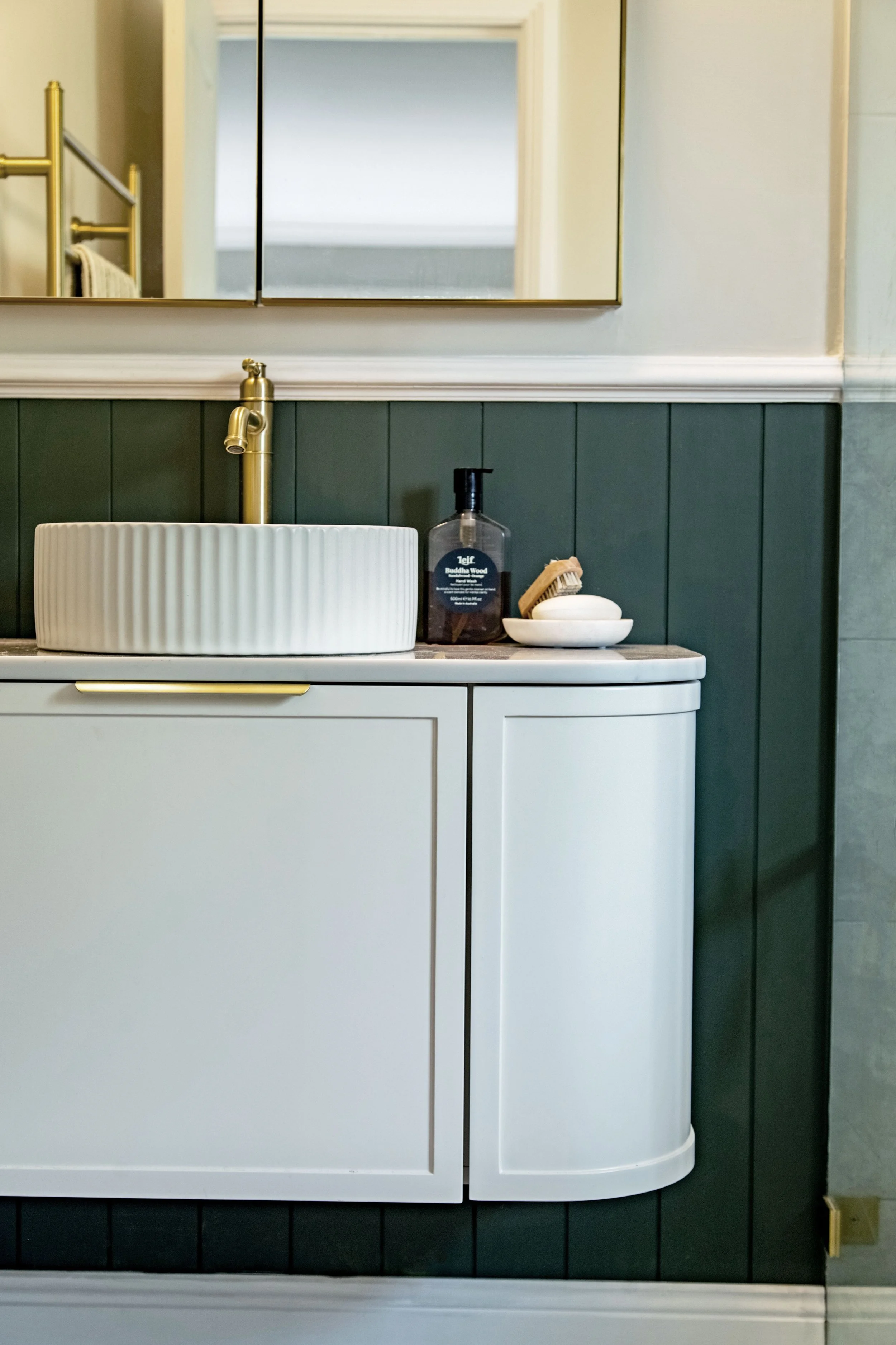 Bathroom vanity with a white vertical ribbed vessel sink, a gold faucet, a soap dispenser, and a tray with soap and brushes. Mirror above the vanity, green paneled wall, white cabinet base, and a door reflected in the mirror.