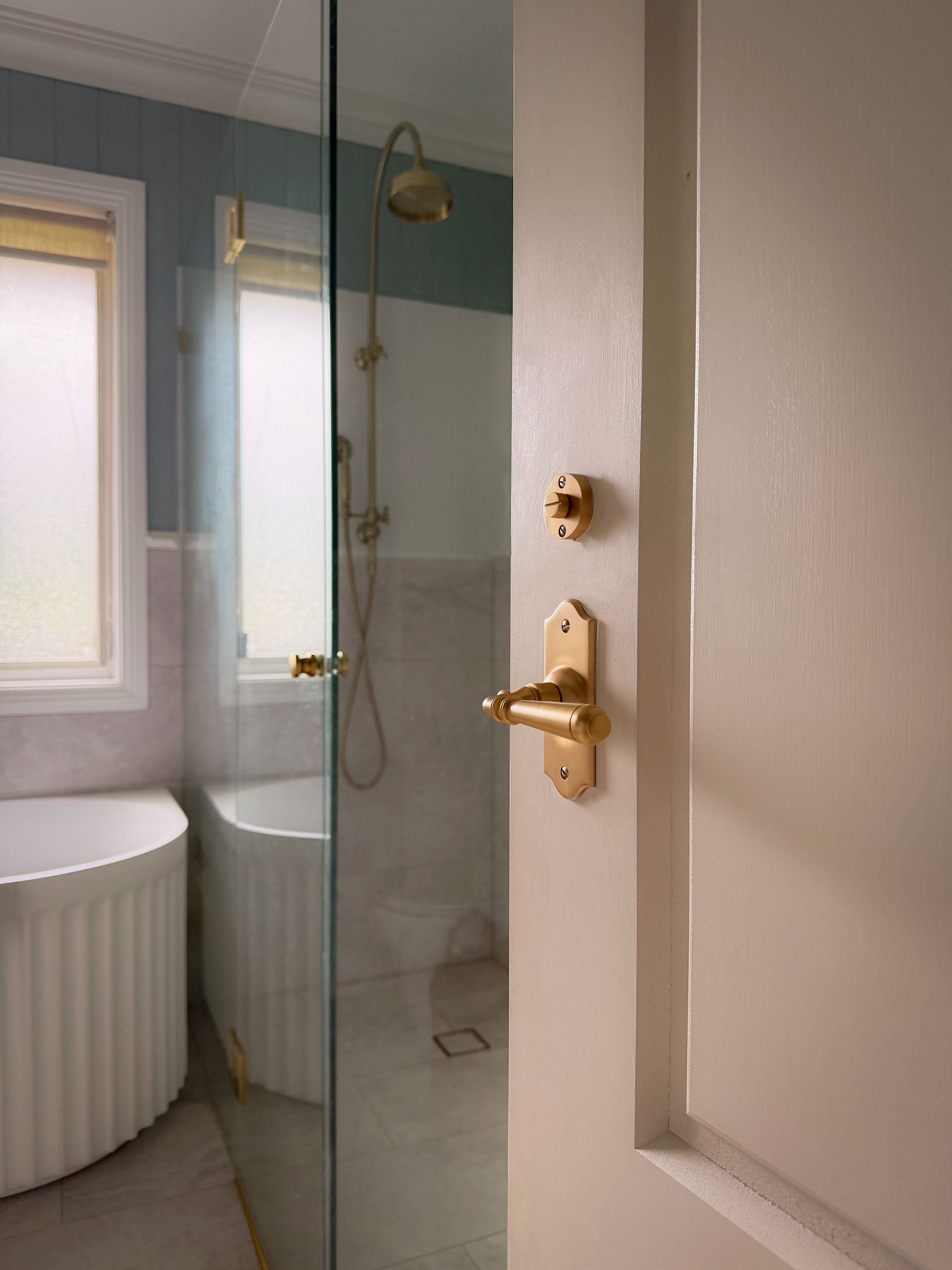 Close-up of a bathroom door with a gold-colored doorknob and lock, partially open revealing a shower area with a showerhead and a glass door, and a window with frosted glass nearby.