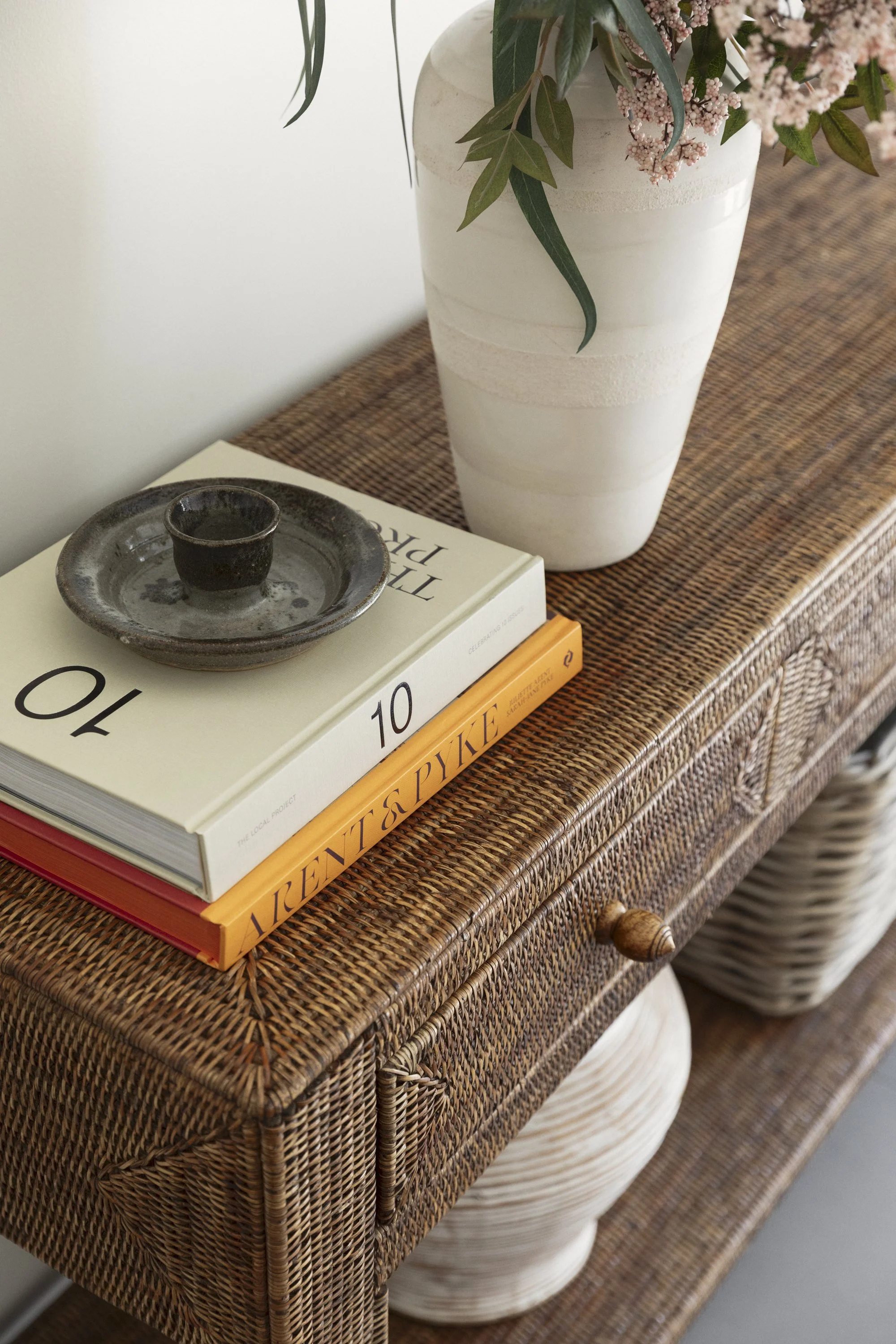 A wicker sideboard with a large white vase containing pink flowers and green leaves, and a small ceramic dish on top of a stack of books.