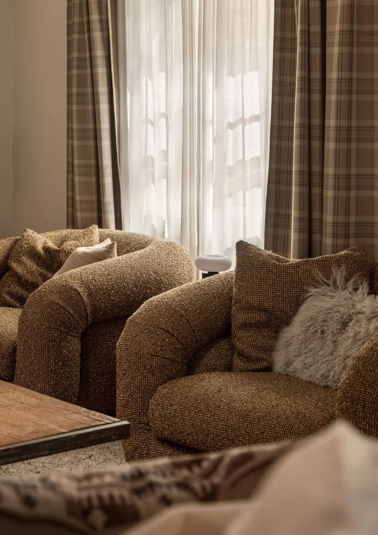 Living room with brown textured armchairs, beige throw pillows, sheer curtains letting in natural light, and a fluffy gray throw pillow.