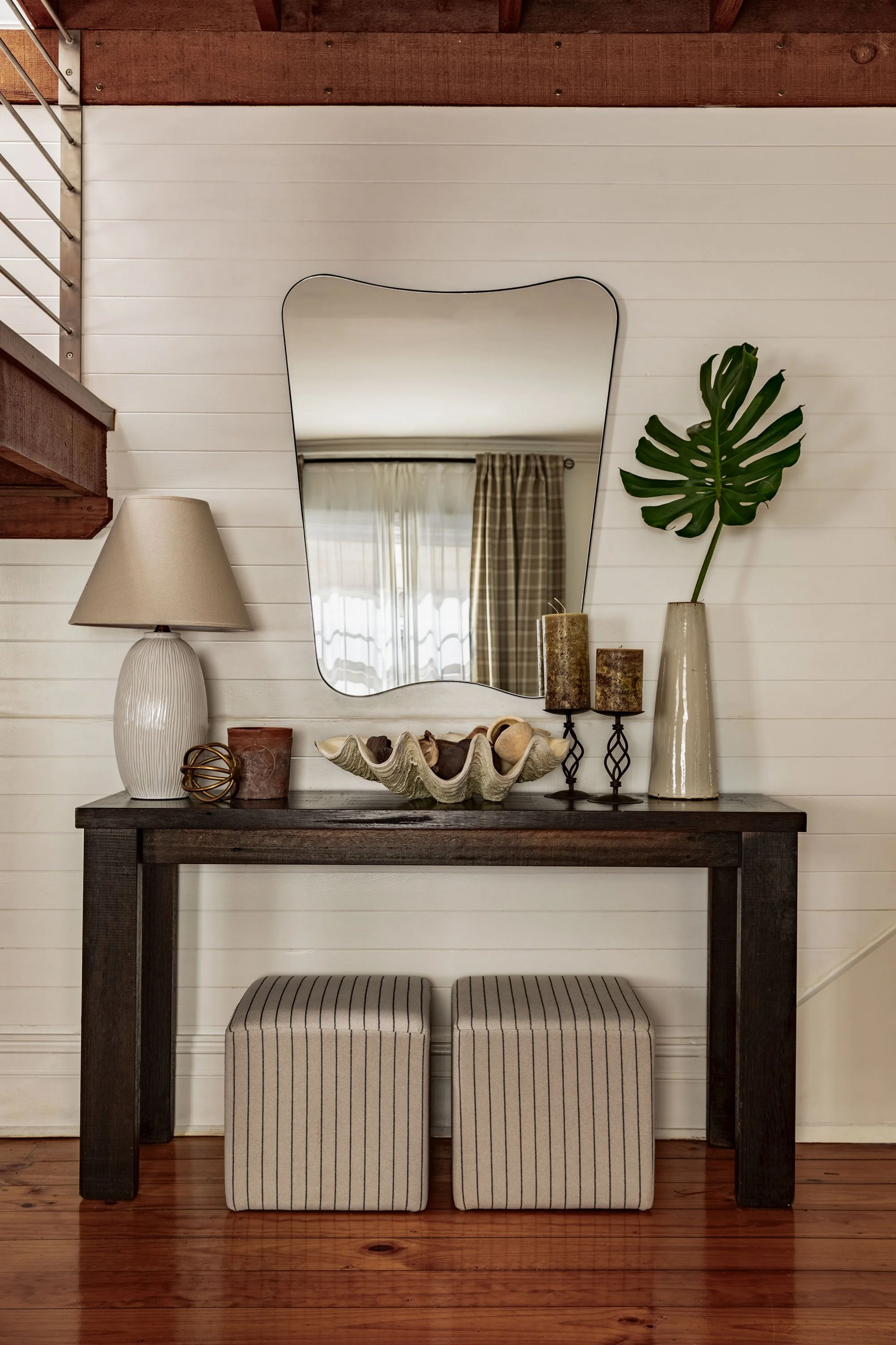 Decorative console table with a mirror, lamp, large leaf in a vase, candles, and shells, set against a white shiplap wall with a window and curtains reflected in the mirror. Two striped ottomans are underneath the table, on a wooden floor.