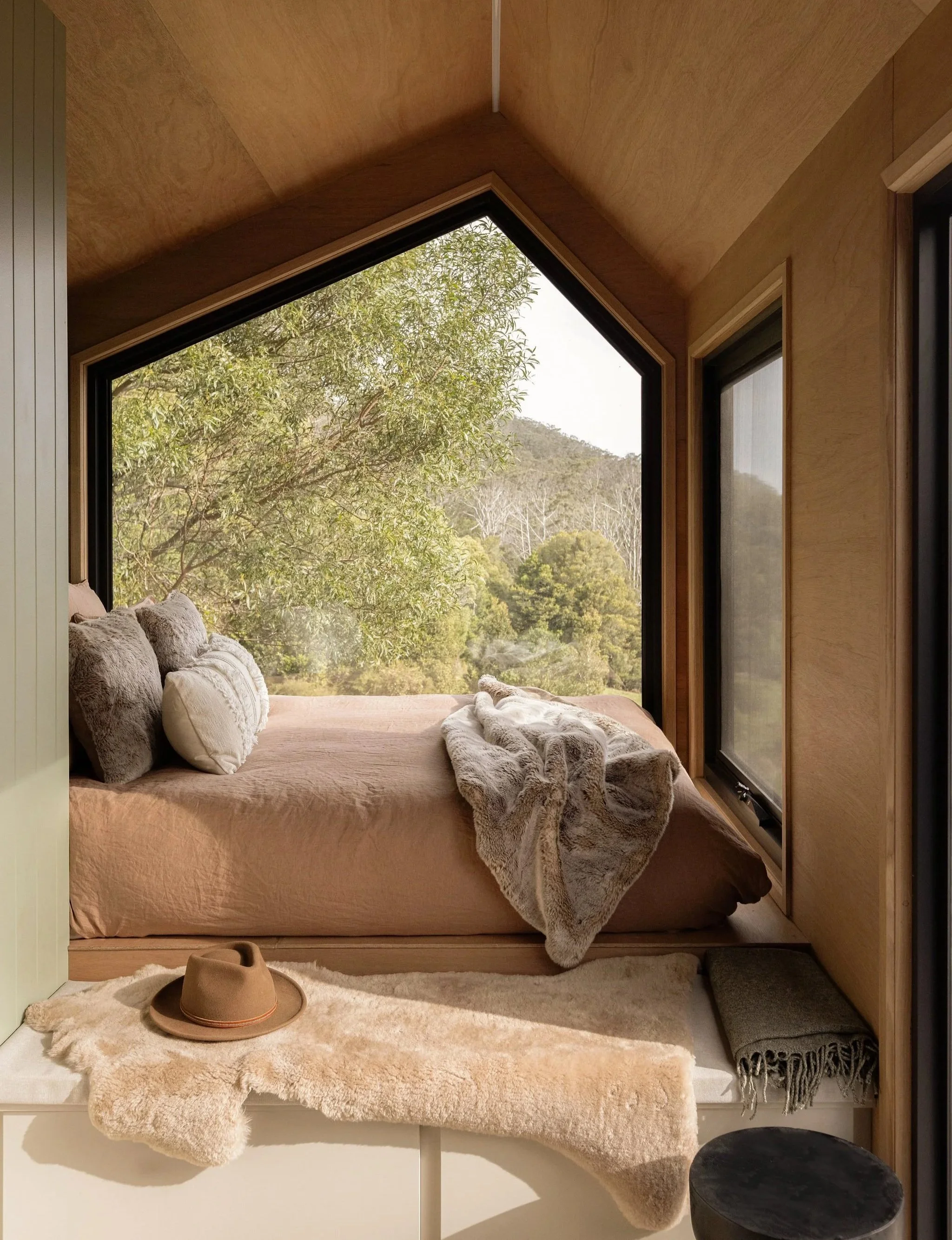 A cozy window nook with a bed, pillows, and a plush blanket, overlooking a lush green landscape with trees and hills.