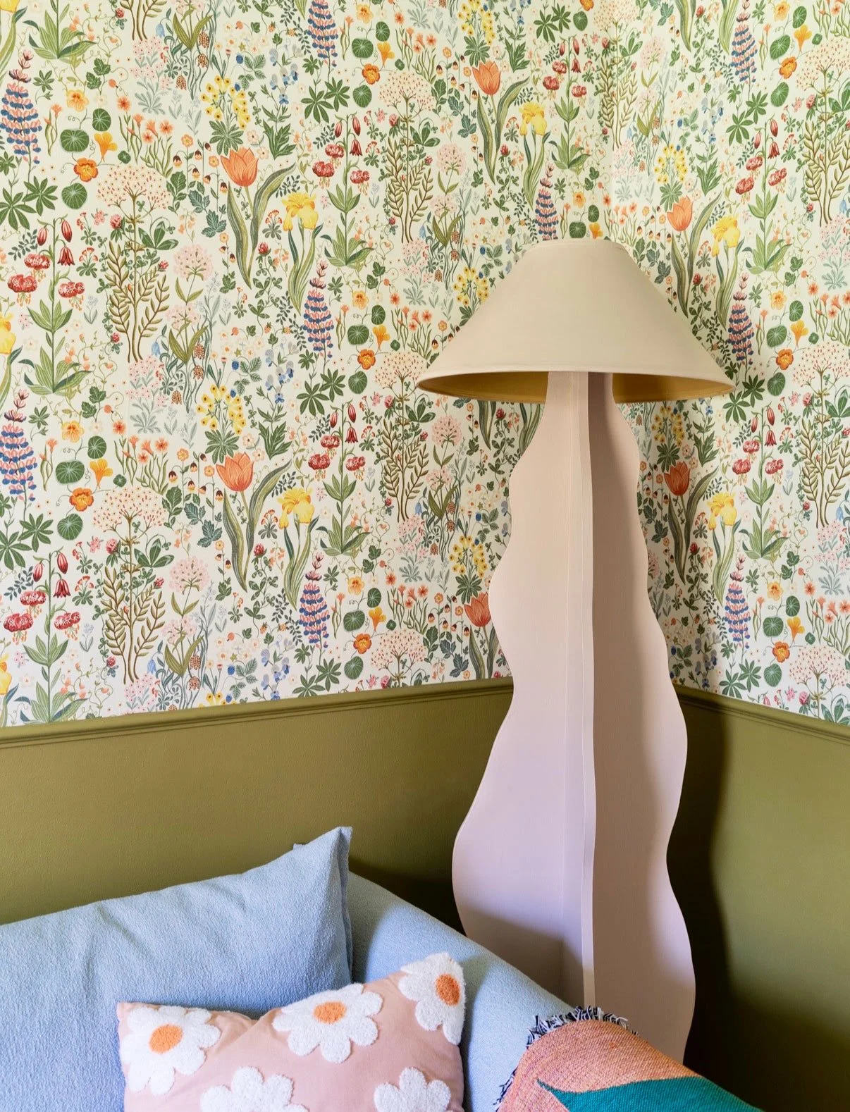 A wall with floral wallpaper, a abstract-shaped white lamp, a bed with blue pillows, and a pink pillow with white daisies and an orange center.