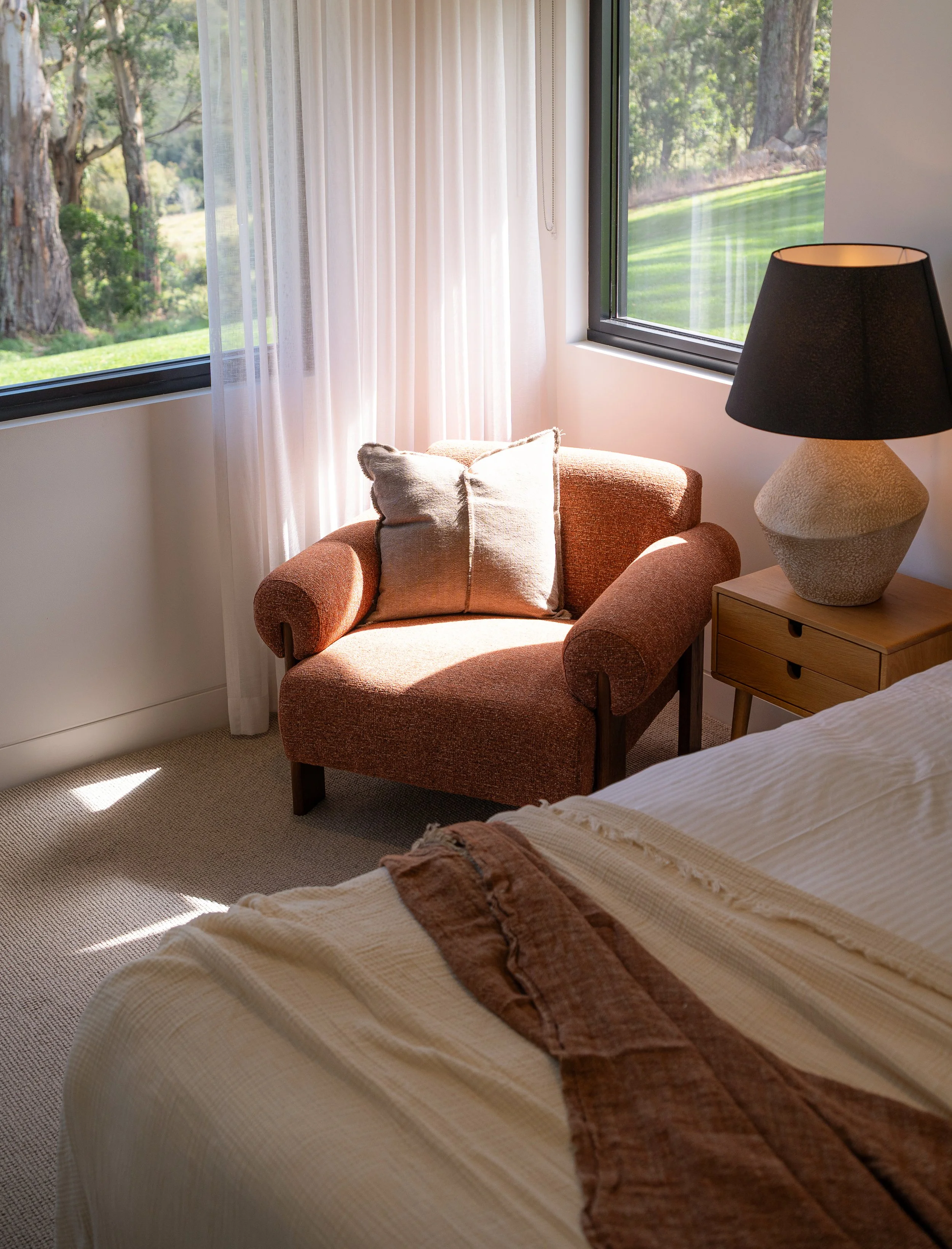 A cozy bedroom corner with a bed covered in a beige checkered bedspread, a rust-colored throw blanket, and a striped pillow. Next to the bed, a wooden nightstand holds a large textured beige and black table lamp. The room has two large windows with s