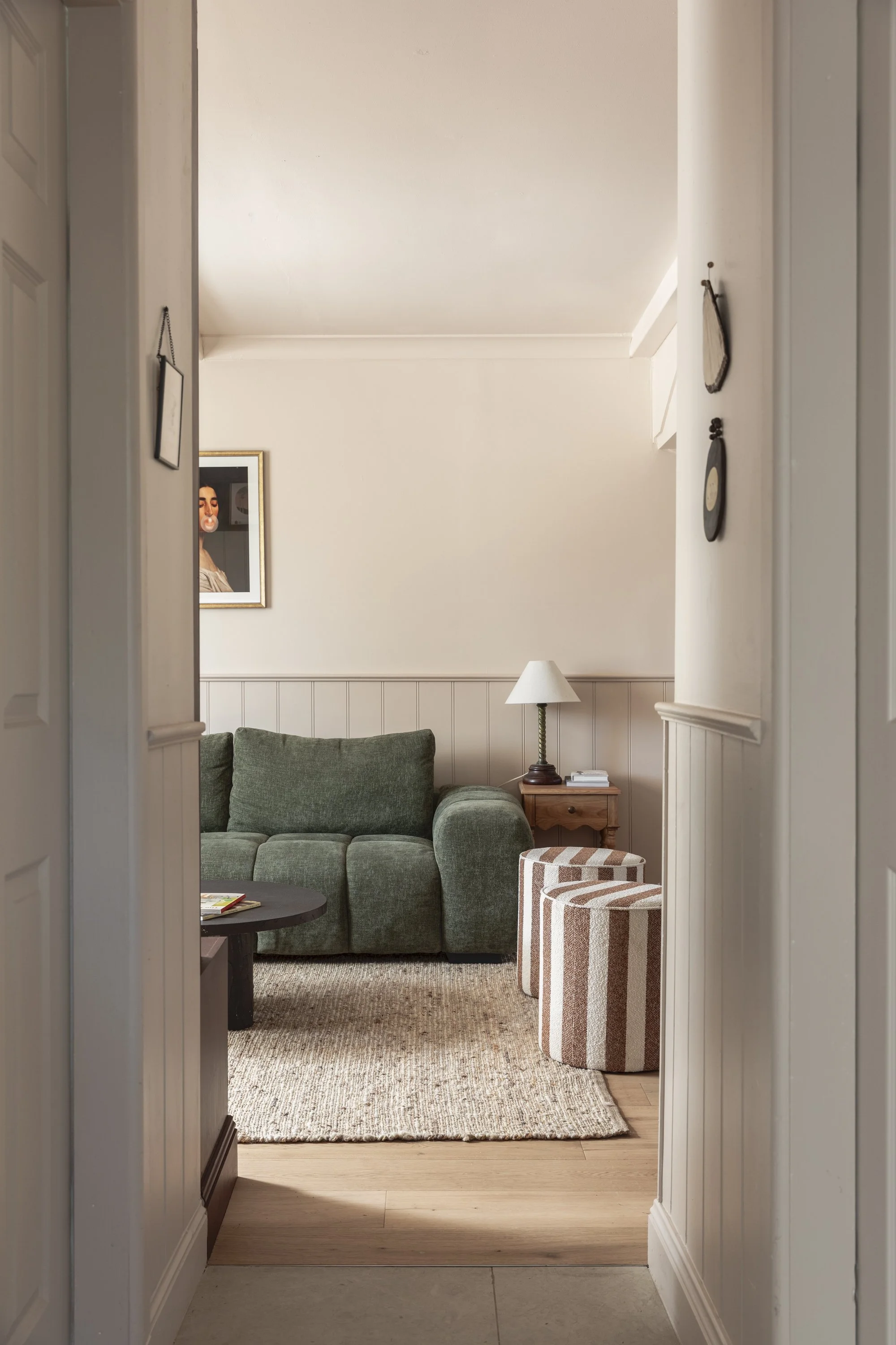 Living room with green sofa, striped ottomans, wooden side table, white wall with framed art, lamp, and a rug.