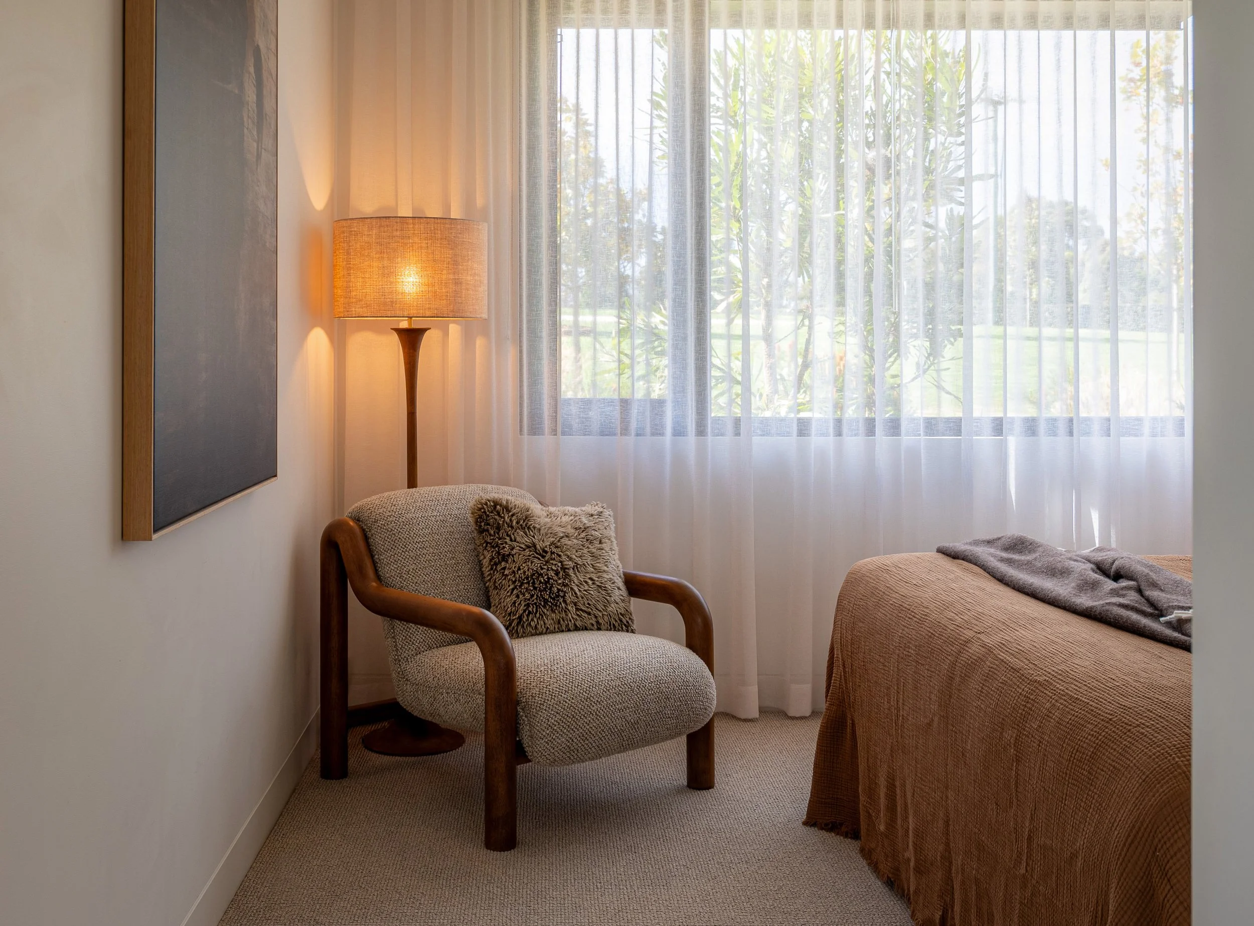 Cozy bedroom corner with a beige upholstered armchair, fluffy throw pillow, and side table, next to a window with sheer curtains letting in natural light.