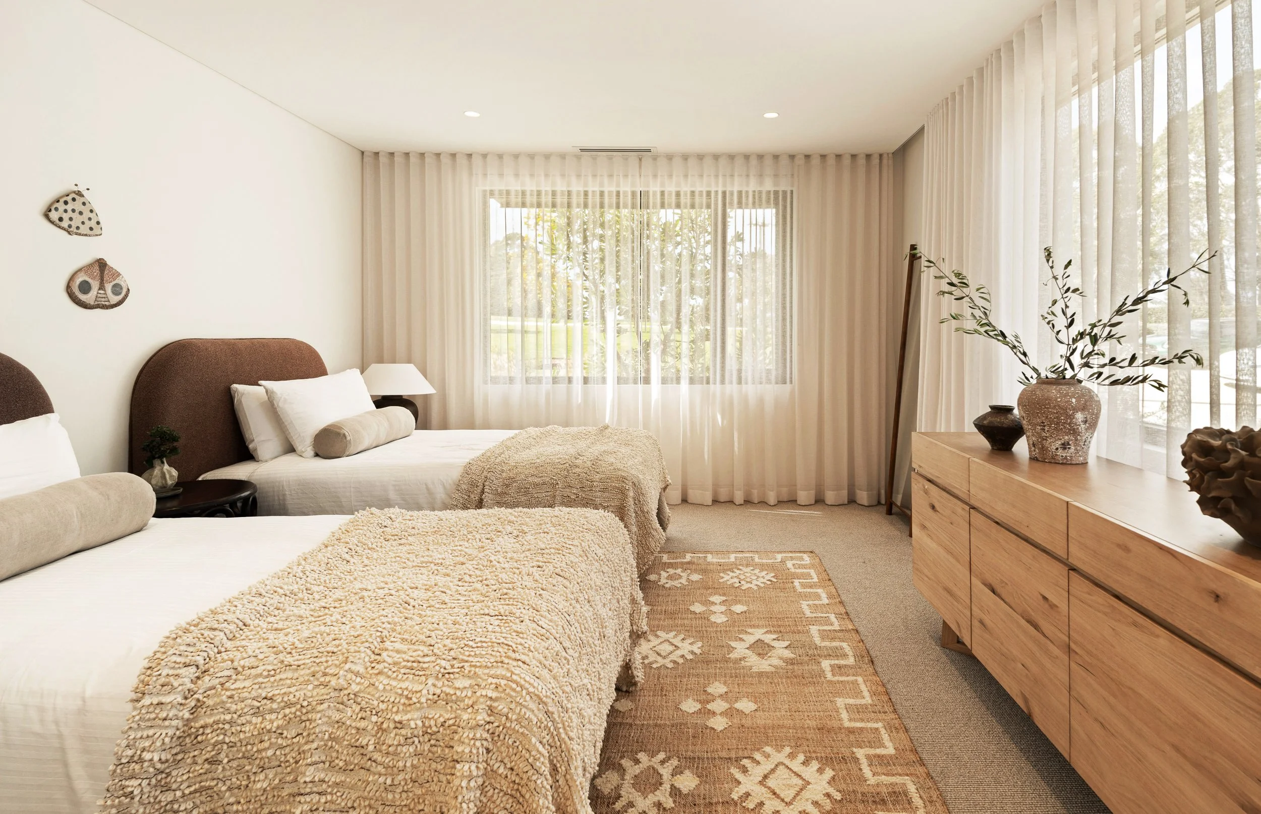 A bedroom with two beds, rust coloured headboards, white bedding, a wooden dresser with vases, a large window with sheer curtains, and wall decorations, including a butterfly and a moth.
