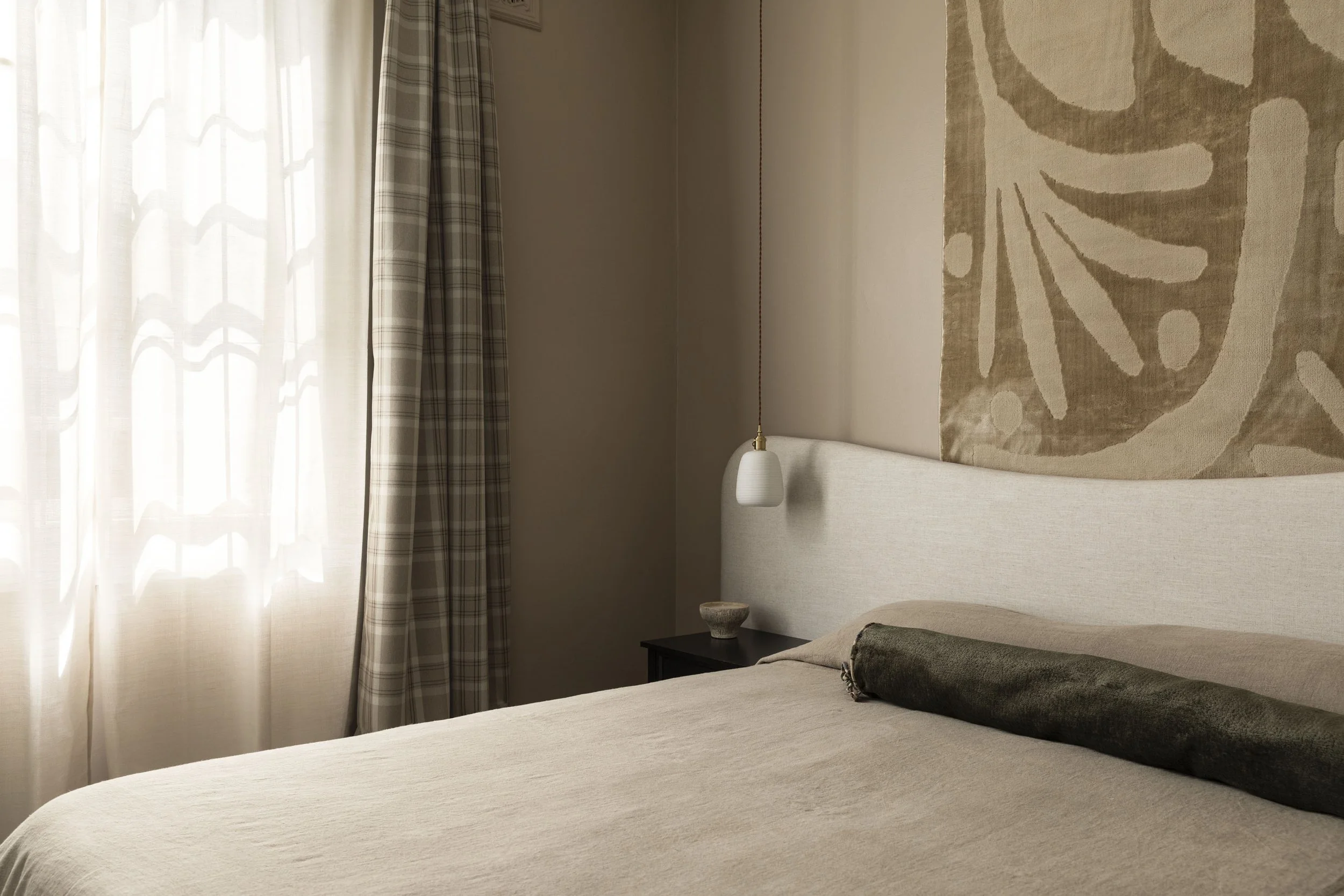 A neatly made bed with a beige bedspread, a dark green bolster pillow, and a cream-colored headboard. A small bedside table with a decorative bowl and a hanging white pendant light are next to the bed. Light filters through sheer curtains beside a patterned curtain on a window.