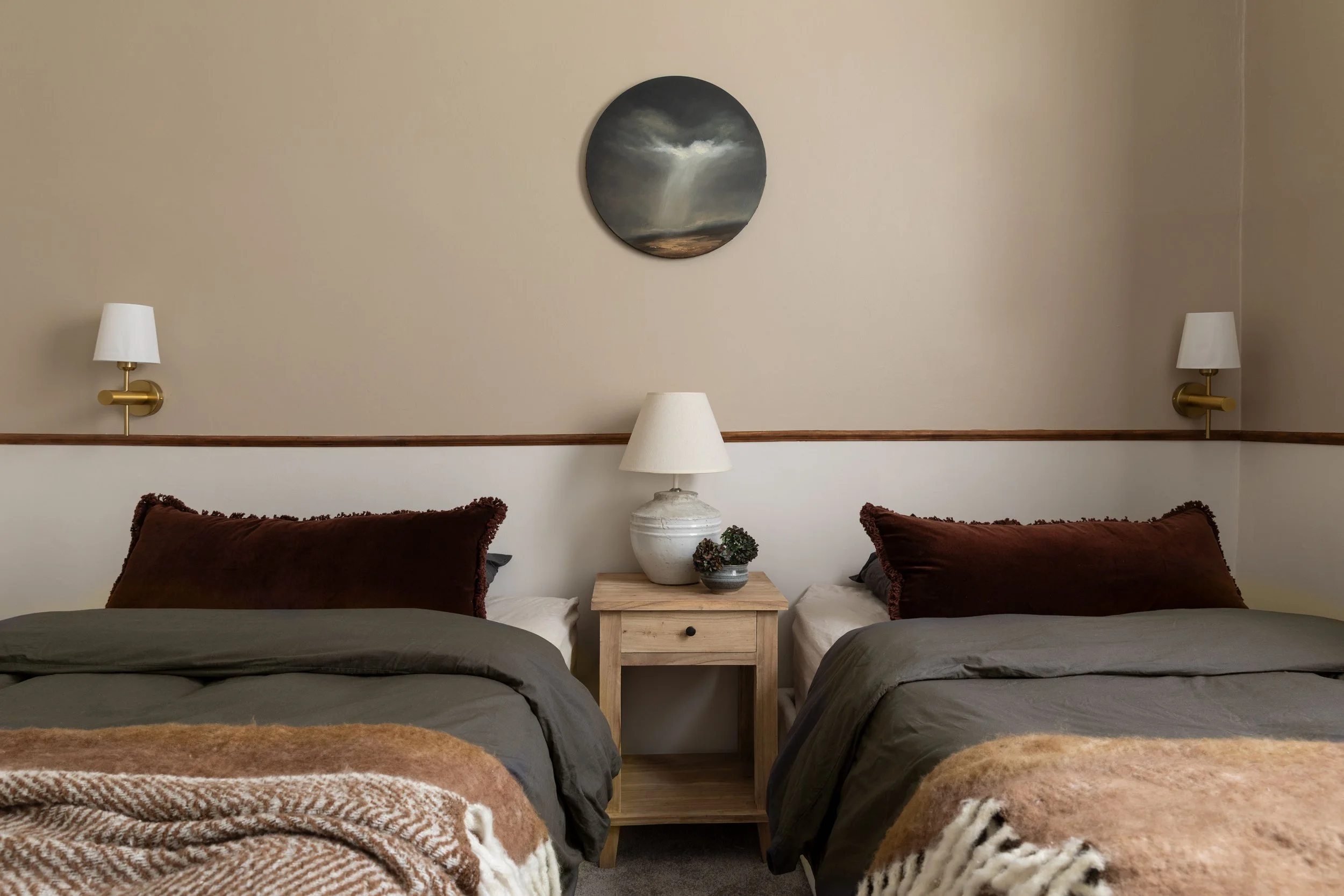 A bedroom with two beds separated by a wooden nightstand, with a lamp and small plants on it. The beds have dark pillows and blankets, and there are two wall-mounted lamps above each bed. A round painting of a stormy sky with a lightning bolt hangs above the headboards.