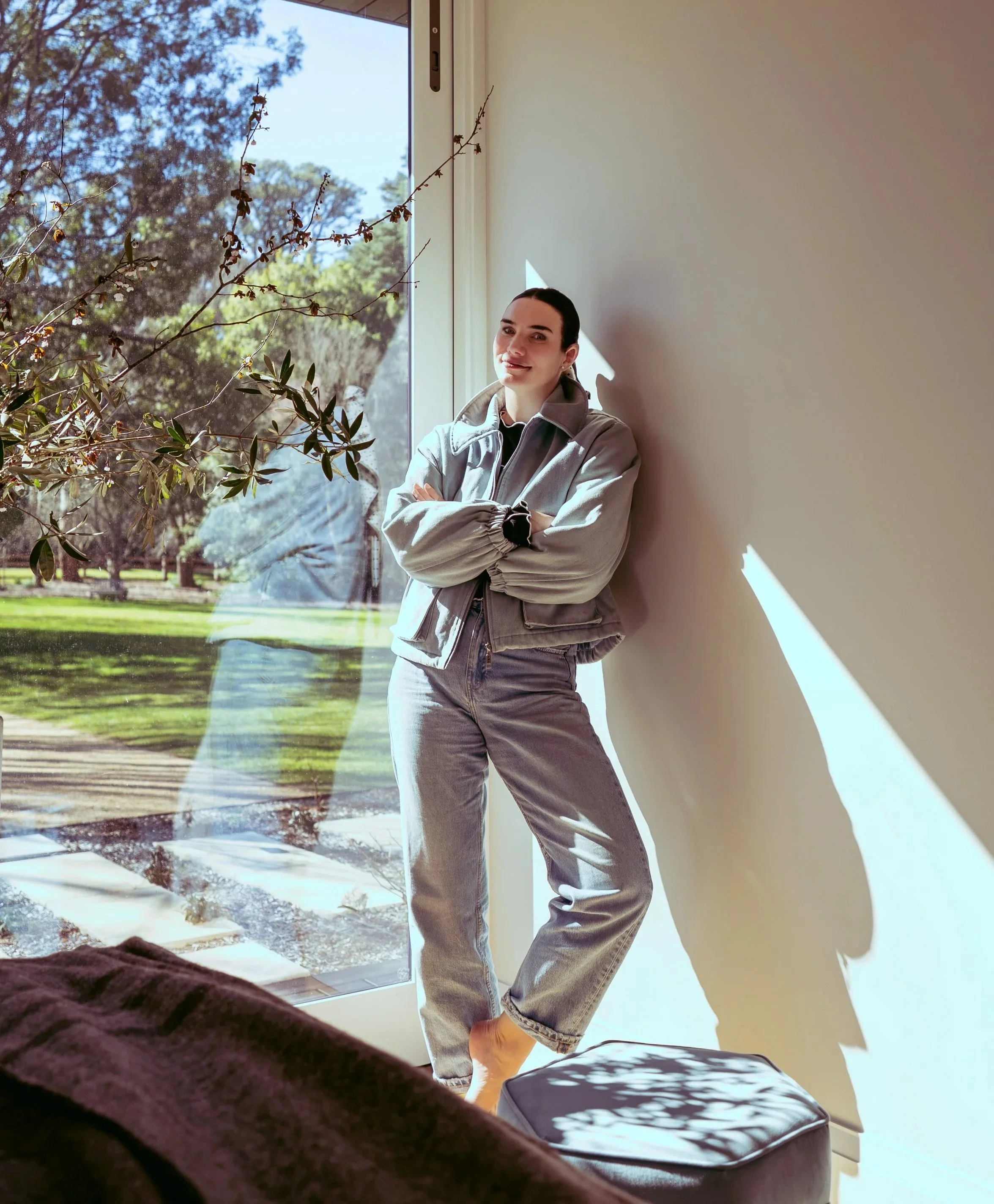 A woman with dark hair pulled back, wearing a light jacket, stands by a large window with her arms crossed, smiling at the camera. Outside the window, trees and a lawn are visible. The indoor setting has sunlight creating shadows on the wall.