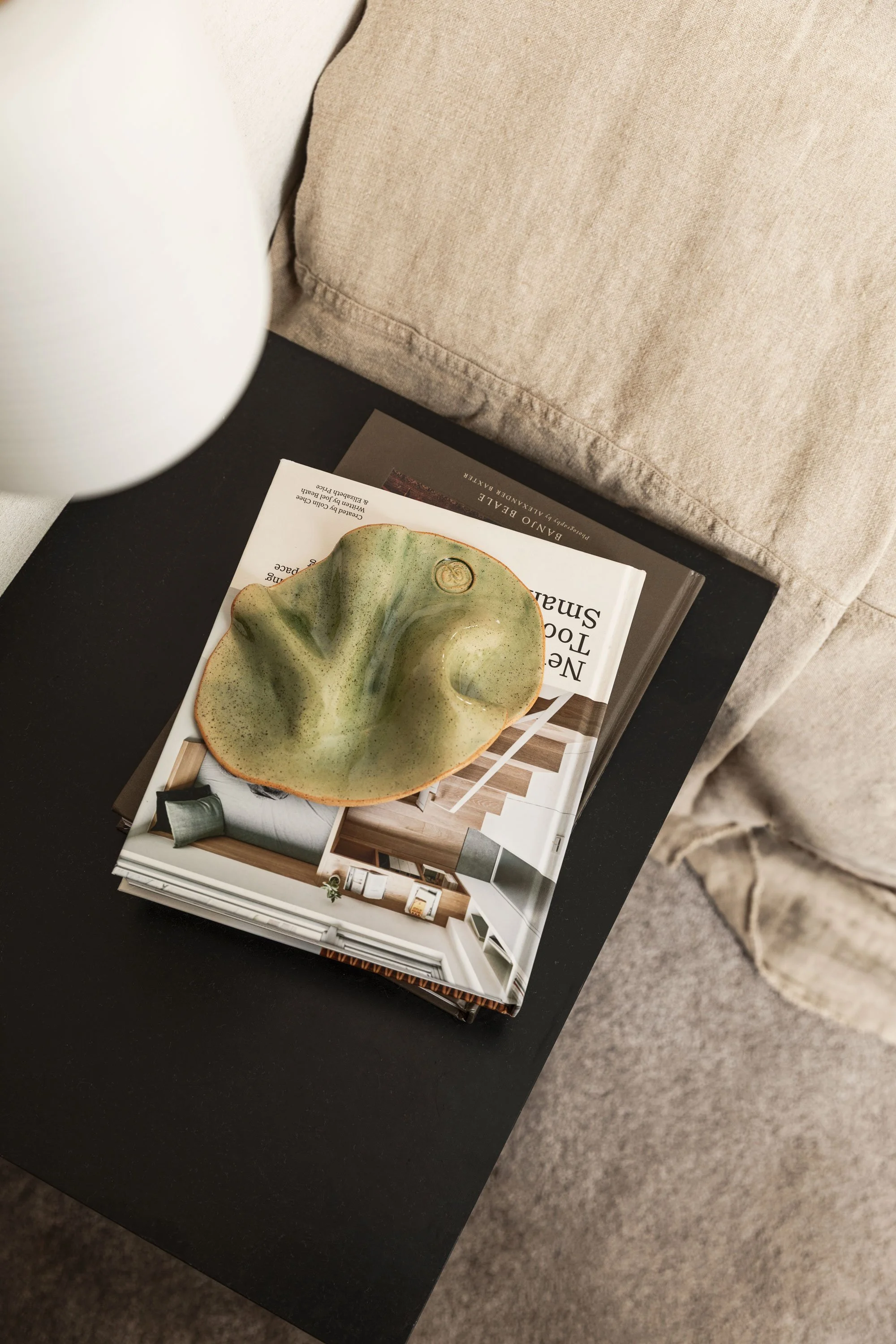 A black side table holding a stack of interior design magazines with a glazed ceramic bowl-shaped decorative piece on top, next to a beige fabric sofa with textured cushions.