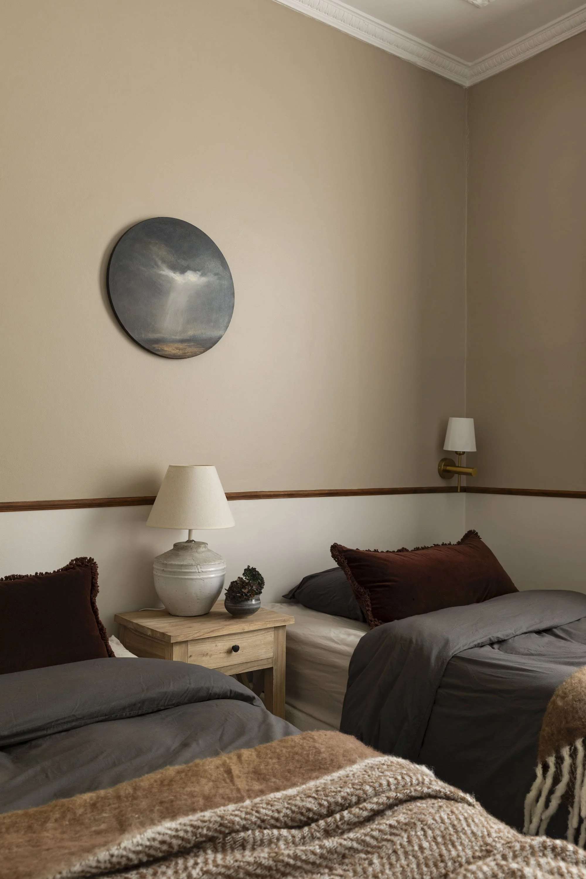 A cozy bedroom with two beds, decorated with dark brown and gray bedding, and a wooden nightstand between them holding a table lamp and a small potted plant. There are wall-mounted lamps on each side of the beds and a round landscape painting above.