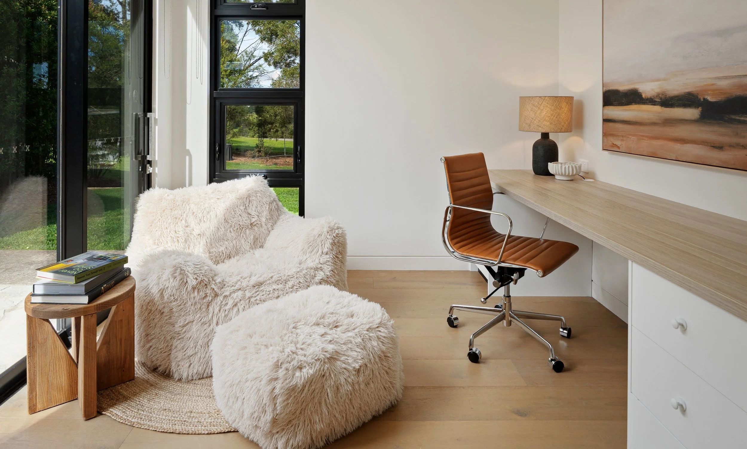 A modern home office with a white furry armchair next to a window, a wooden side table with books, a desk with a tan leather swivel chair, a table lamp, a decorative bowl, and a large landscape painting on the wall.