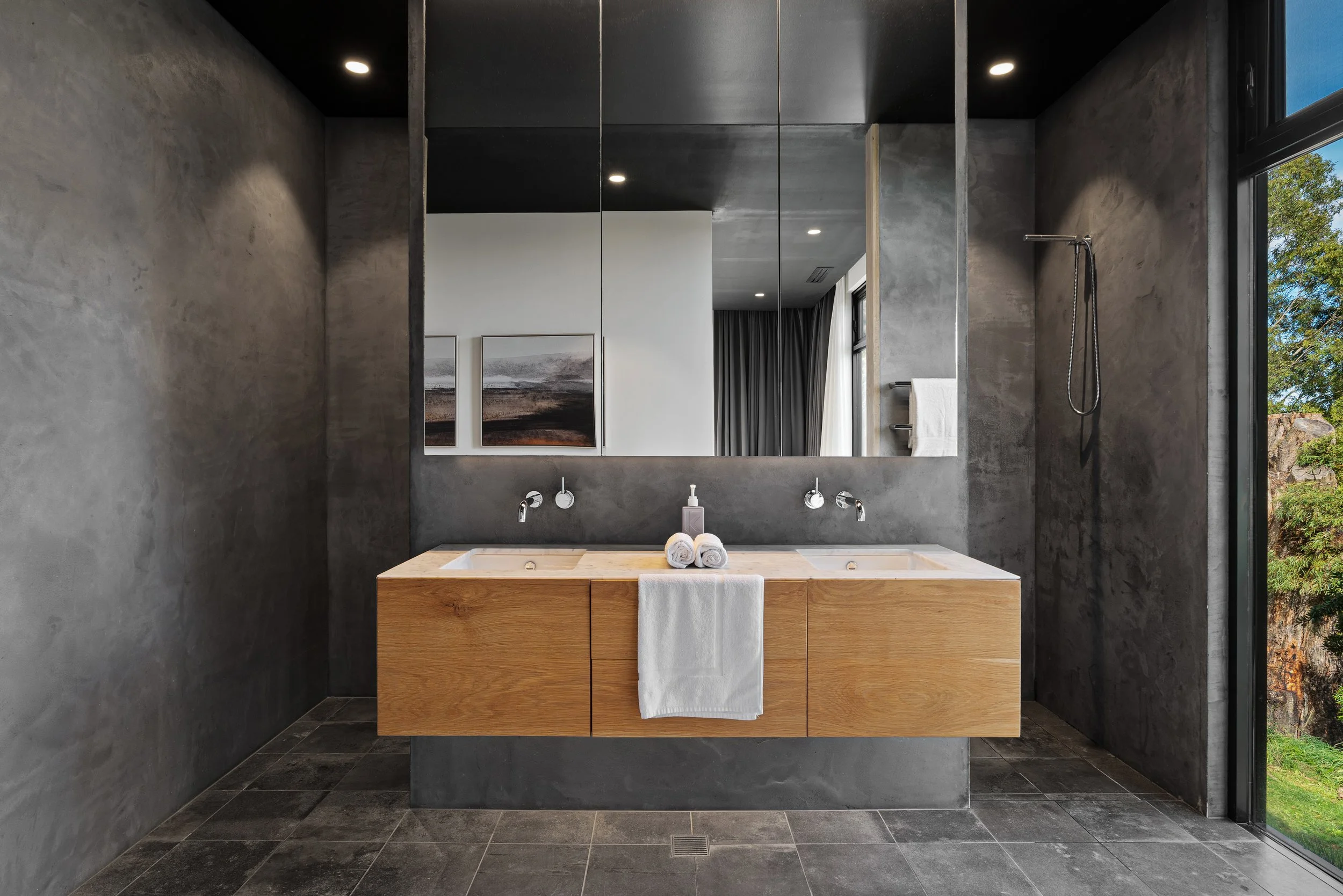 Modern bathroom with dark gray textured walls, black ceiling, and black tiled floor. Features a floating wooden vanity with two integrated sinks, soap dispenser, and towels. Large mirror above the vanity reflects a white wall with two framed landscape paintings, a window with gray curtains, and a bathroom shower with a handheld showerhead on the right side. Natural light from the window illuminates the space.