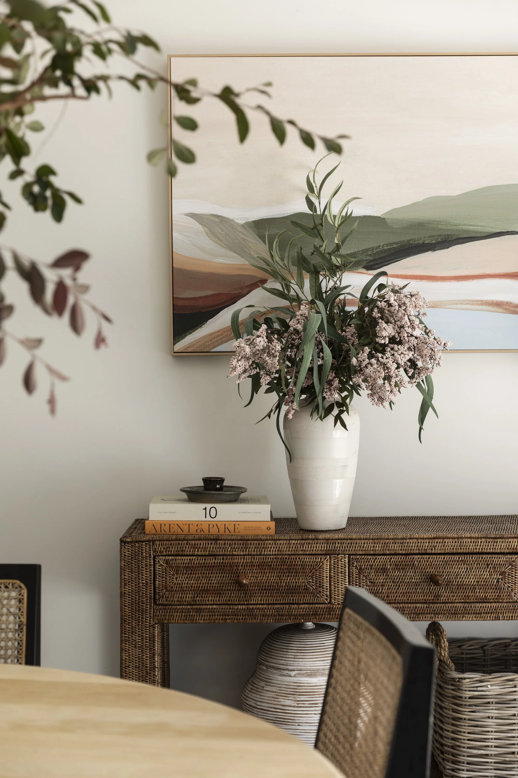 A living room corner with a wicker sideboard topped by a large white vase with pink flowers and greenery, a stack of books, and a black dish, with a colorful abstract landscape painting on the wall.