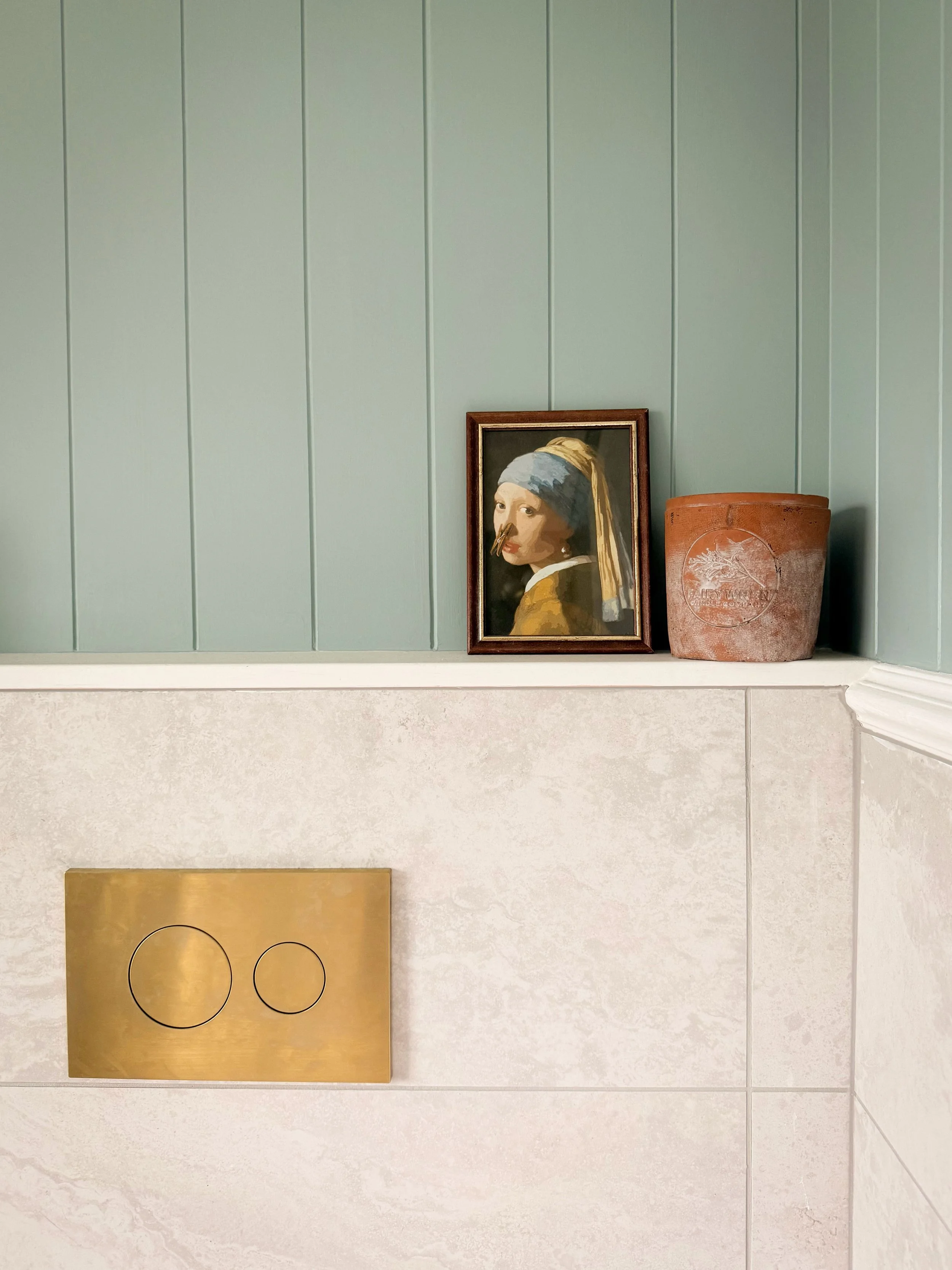 A framed replica of Vermeer’s "Girl with a Pearl Earring" on a light green wall, next to a round terracotta planter with white markings, on a white ledge above a beige textured tile wall with a gold flush plate featuring two round buttons.
