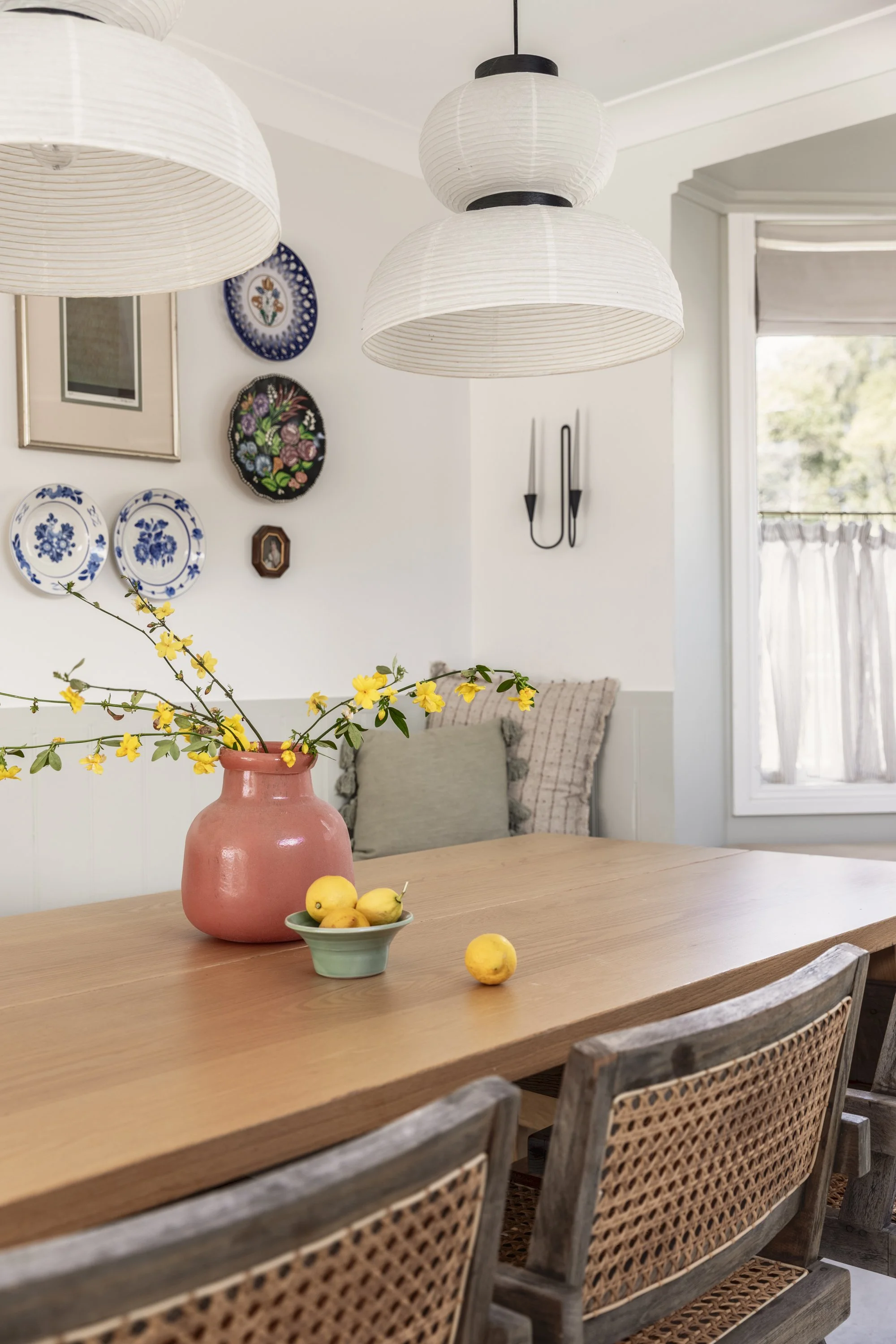 A cozy dining room with a wooden table decorated with a pink vase holding yellow blossoms, a small dish of lemons, and a few lemons scattered on the table. Overhead, there are three white paper lantern pendant lights. The wall features decorative plates and a framed picture, and there's a window with natural light coming in.