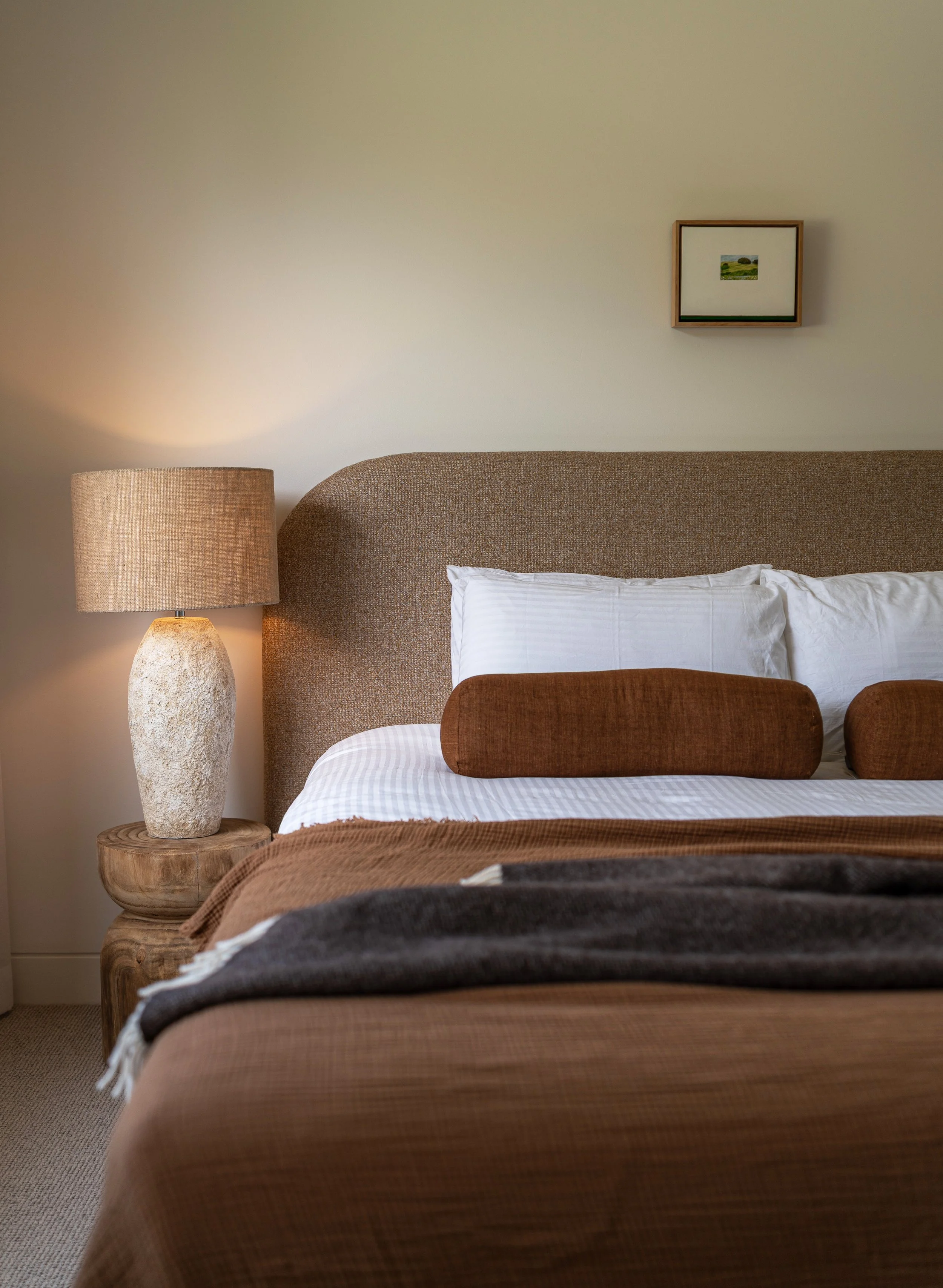A cozy bedroom with a bed, a textured brown headboard, white pillows, and a brown bedspread. Next to the bed is a round, rustic wooden side table with a textured ceramic lamp. On the wall above the bed hangs a small framed landscape painting.