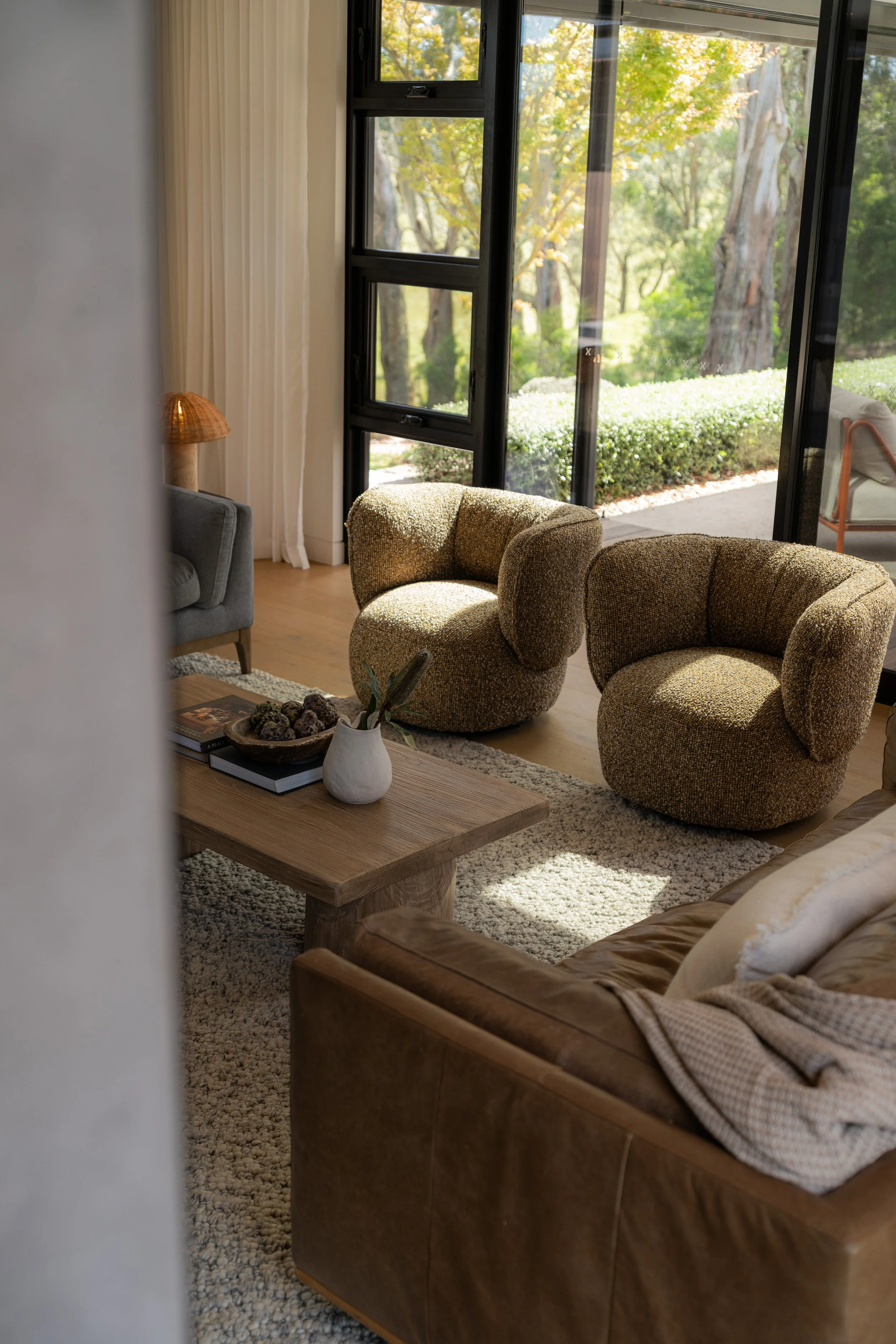 Living room interior with large window view of trees outdoors, brown leather sofa with pillows, and two vintage armchairs near a coffee table with books, a ceramic vase, and a tray of decorative objects.