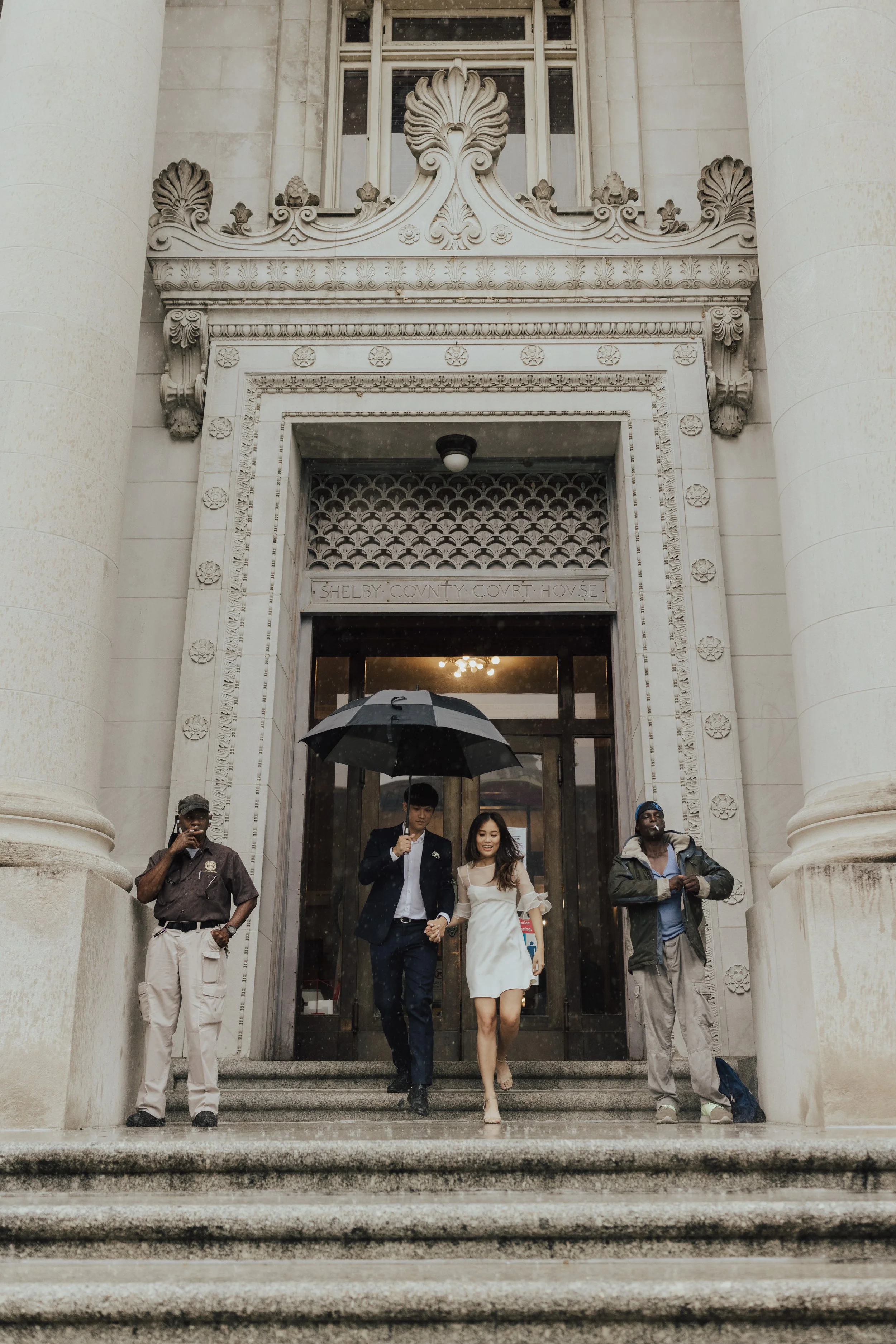 People walking down steps outside stone courthouse building with ornate architectural details, one holding an umbrella.