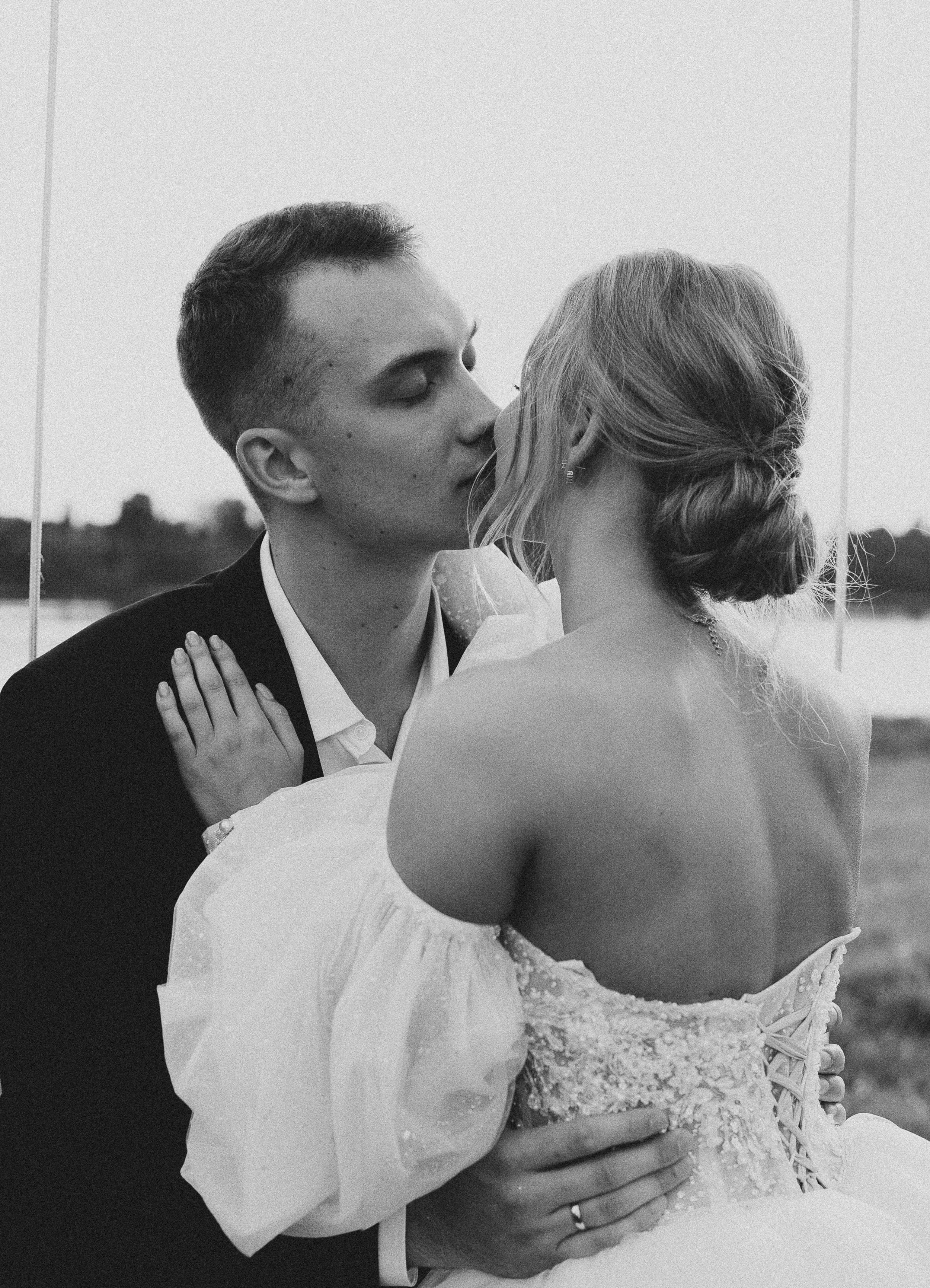 A black-and-white photo of a couple in wedding attire sharing a kiss outdoors, with water and trees in the background.