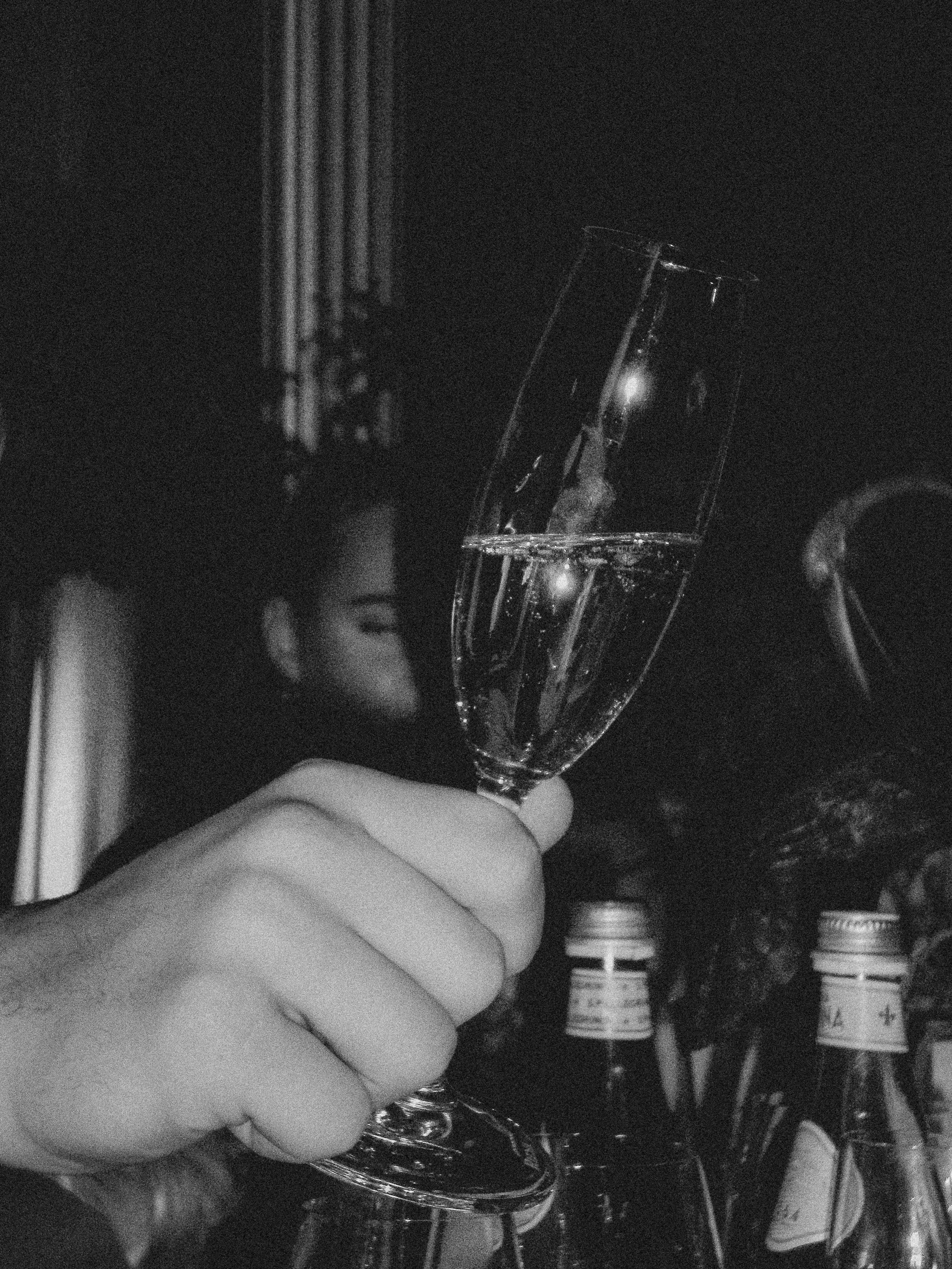 A hand holding a champagne flute filled with sparkling wine, with bottles in the background, in a dark setting.