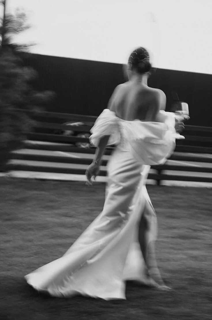 A woman in an elegant off-shoulder satin gown, walking outdoors with a glass of wine, blurred background featuring a fence and trees in black and white.