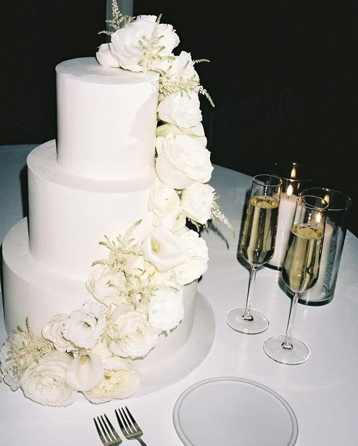 Three-tiered white wedding cake decorated with white flowers and greenery, accompanied by two glasses of champagne and lit candles on a white tablecloth.
