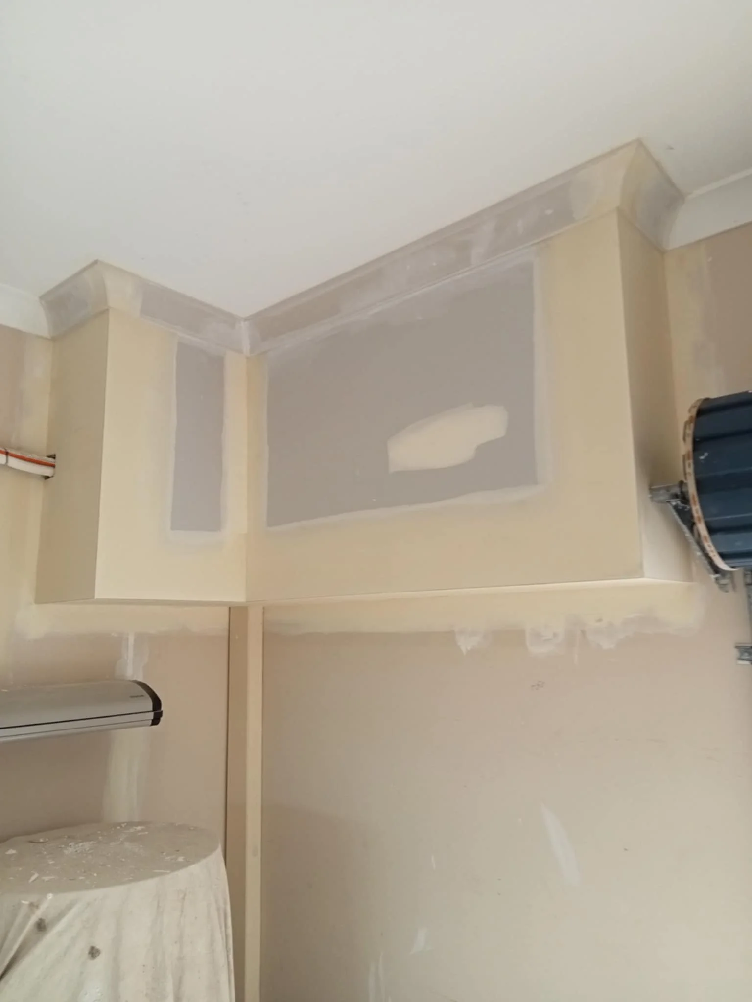 Drywall corner with finished plaster and paint in a room under renovation, featuring a covered object and exposed ceiling.