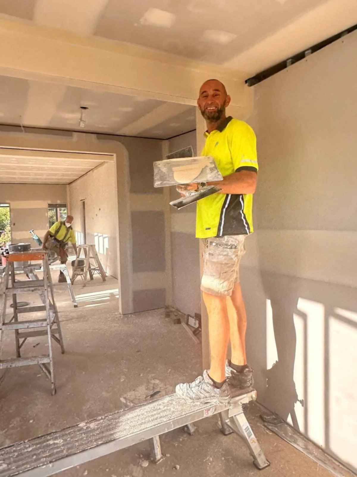 Kurt David, owner and Construction worker standing on scaffold, holding plastering tools inside a partially finished room with drywall.