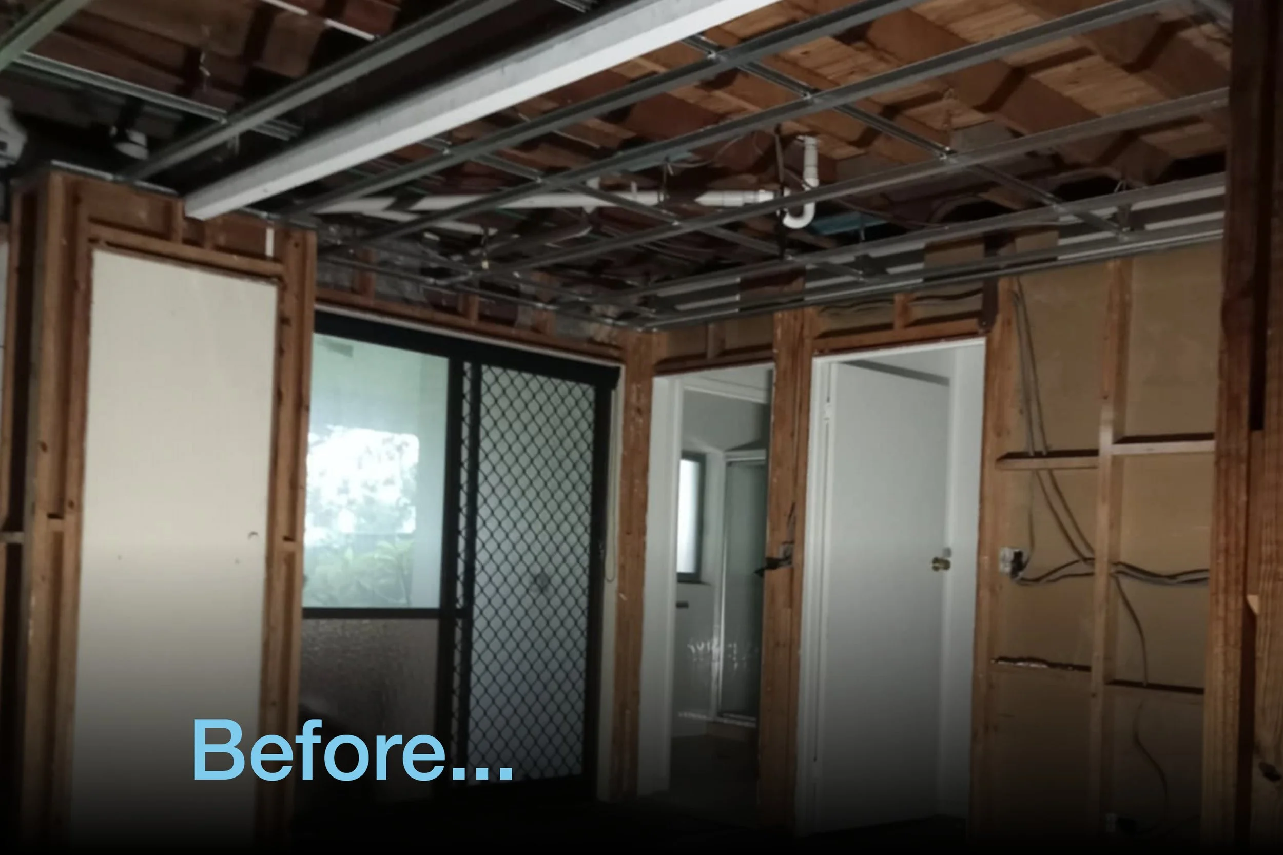 Interior of a room under renovation with exposed wooden frames, visible ceiling pipes, and a sliding glass door with a security screen."}