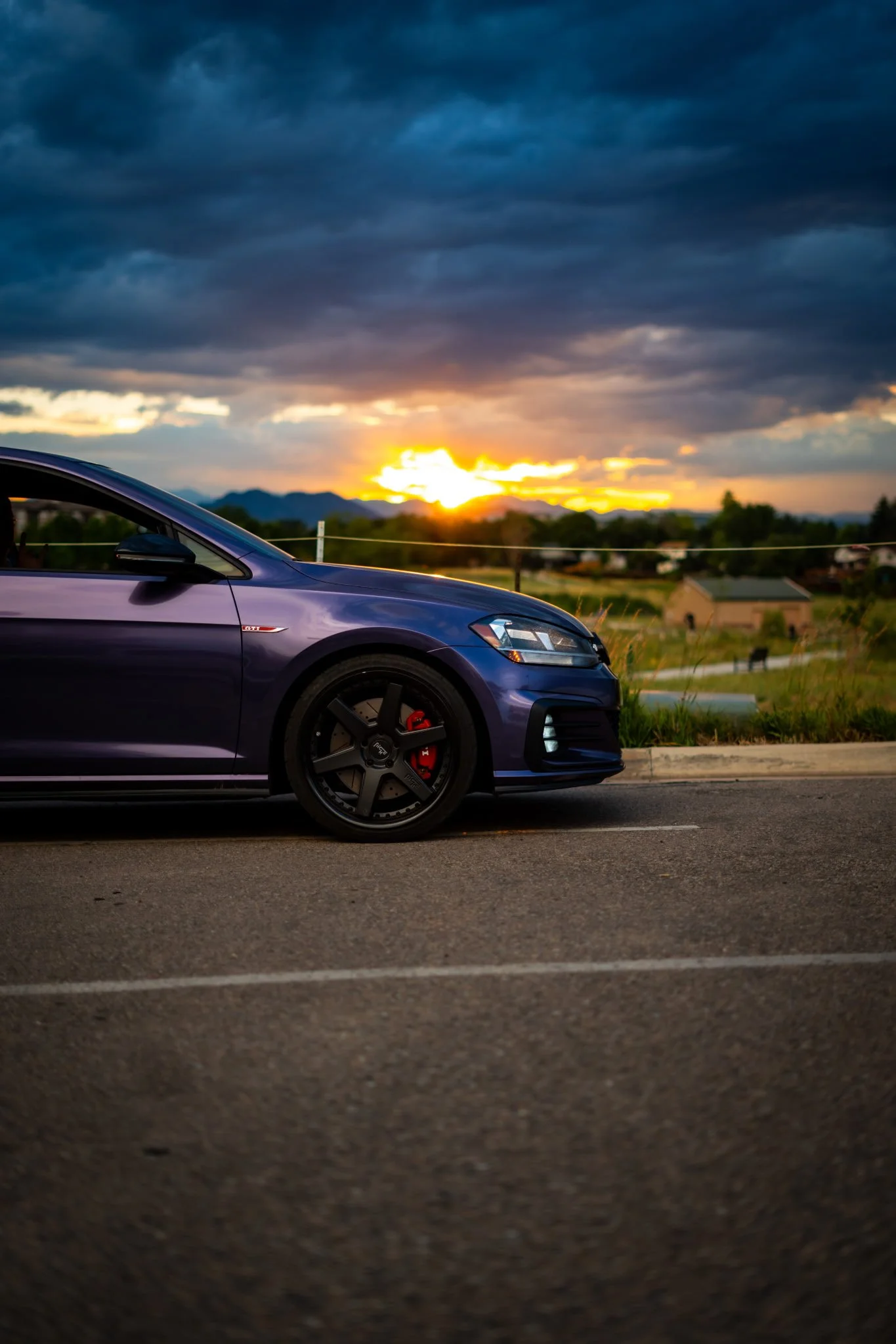 A purple hatchback car parked near a lake with trees along the shore and a cloudy sky in the background.