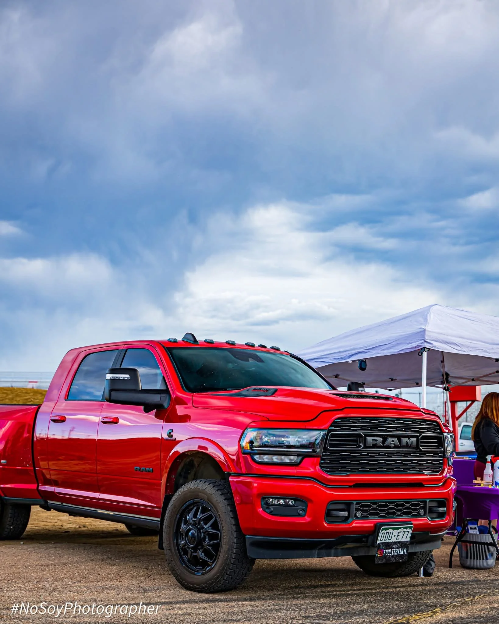 Red RAM pickup truck parked on a dirt roadside near a body of water with mountains in the background and blue sky with clouds.