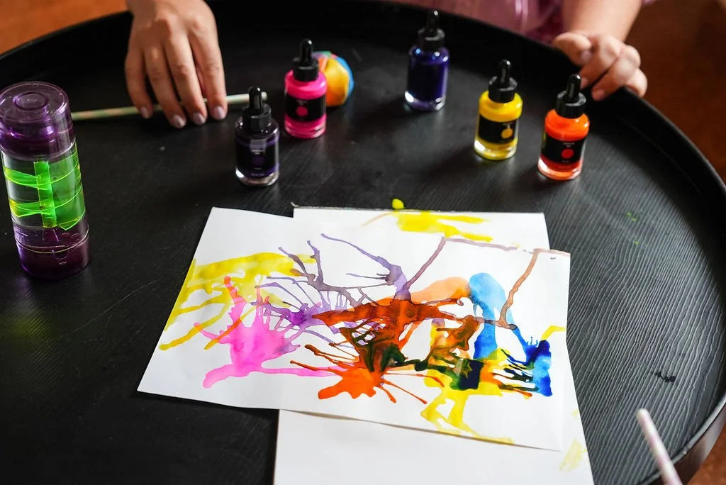 A child's abstract watercolor painting surrounded by bottles of ink and a tray with additional ink bottles on a black surface, the artwork was created as part of a session at the Flourish Collective.