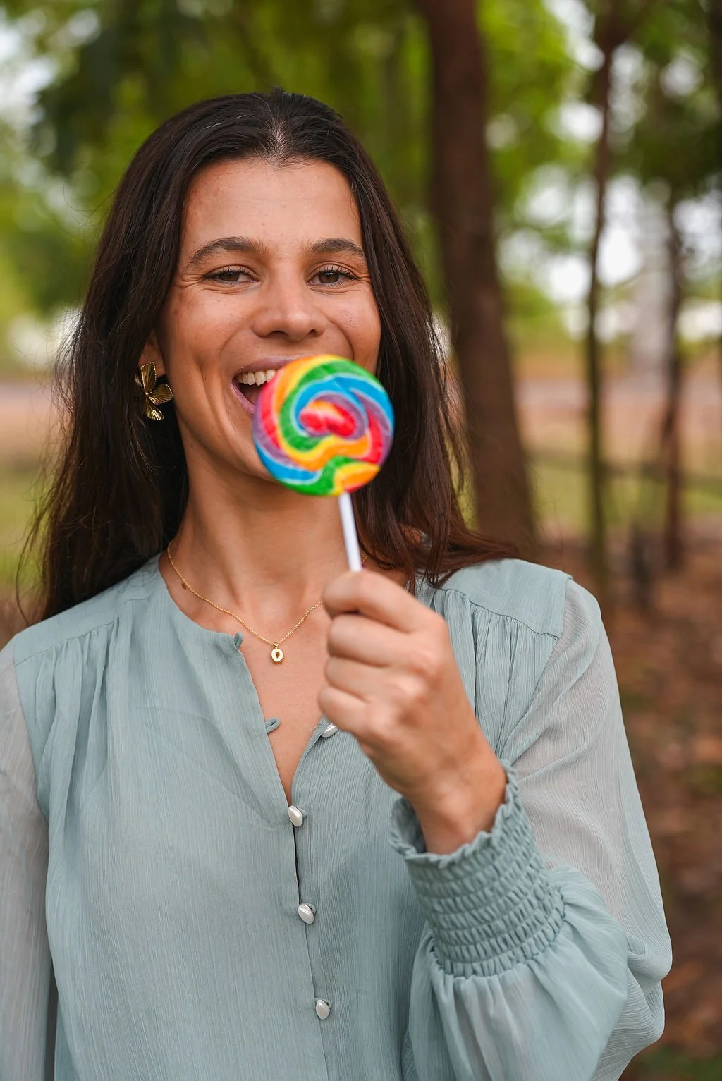 Smiling woman holding a colorful rainbow lollipop outdoors with green trees in the background.