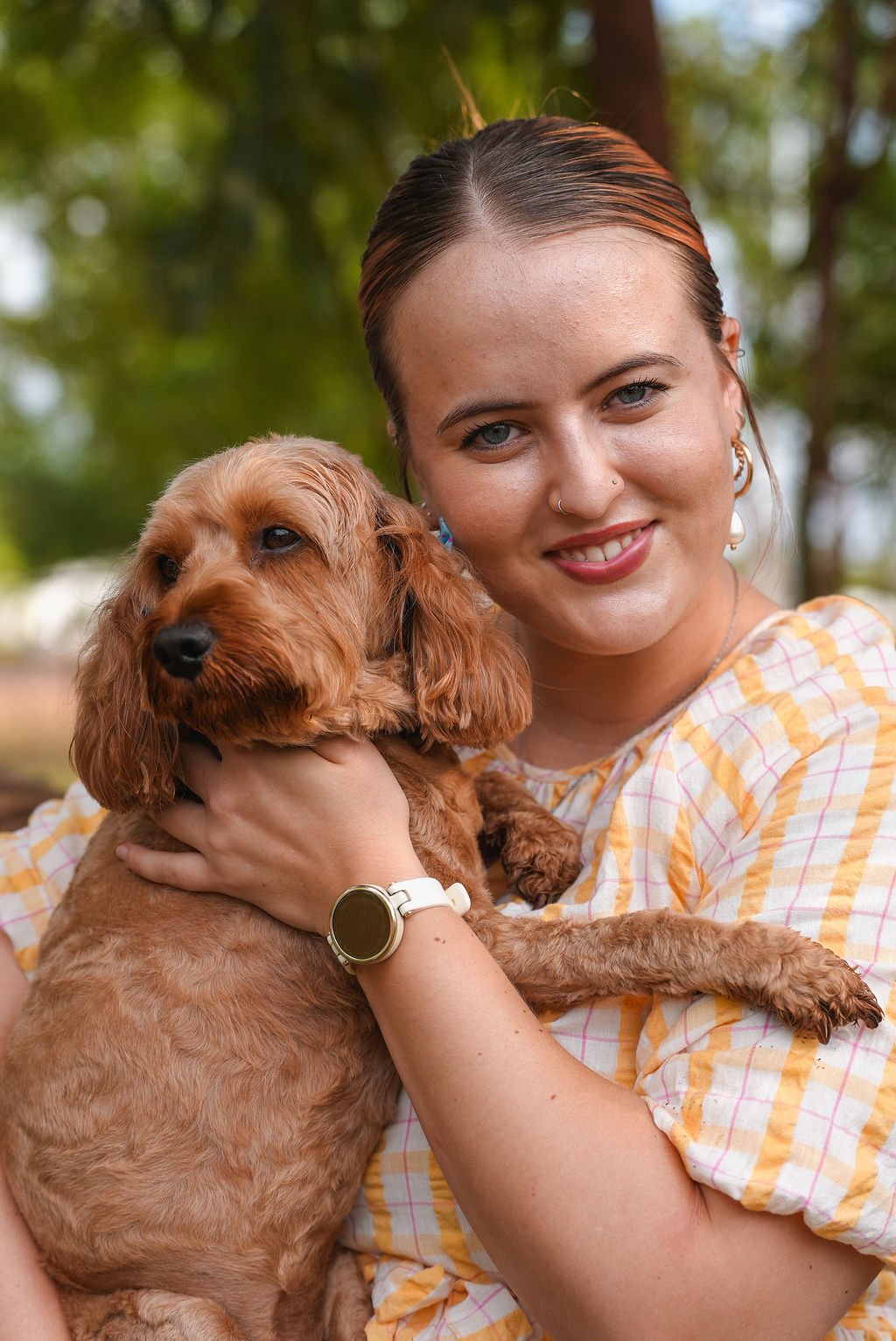 A woman with long hair, blue eyes, and piercings, holding a brown dog outdoors with green trees in the background, smiling at the camera.