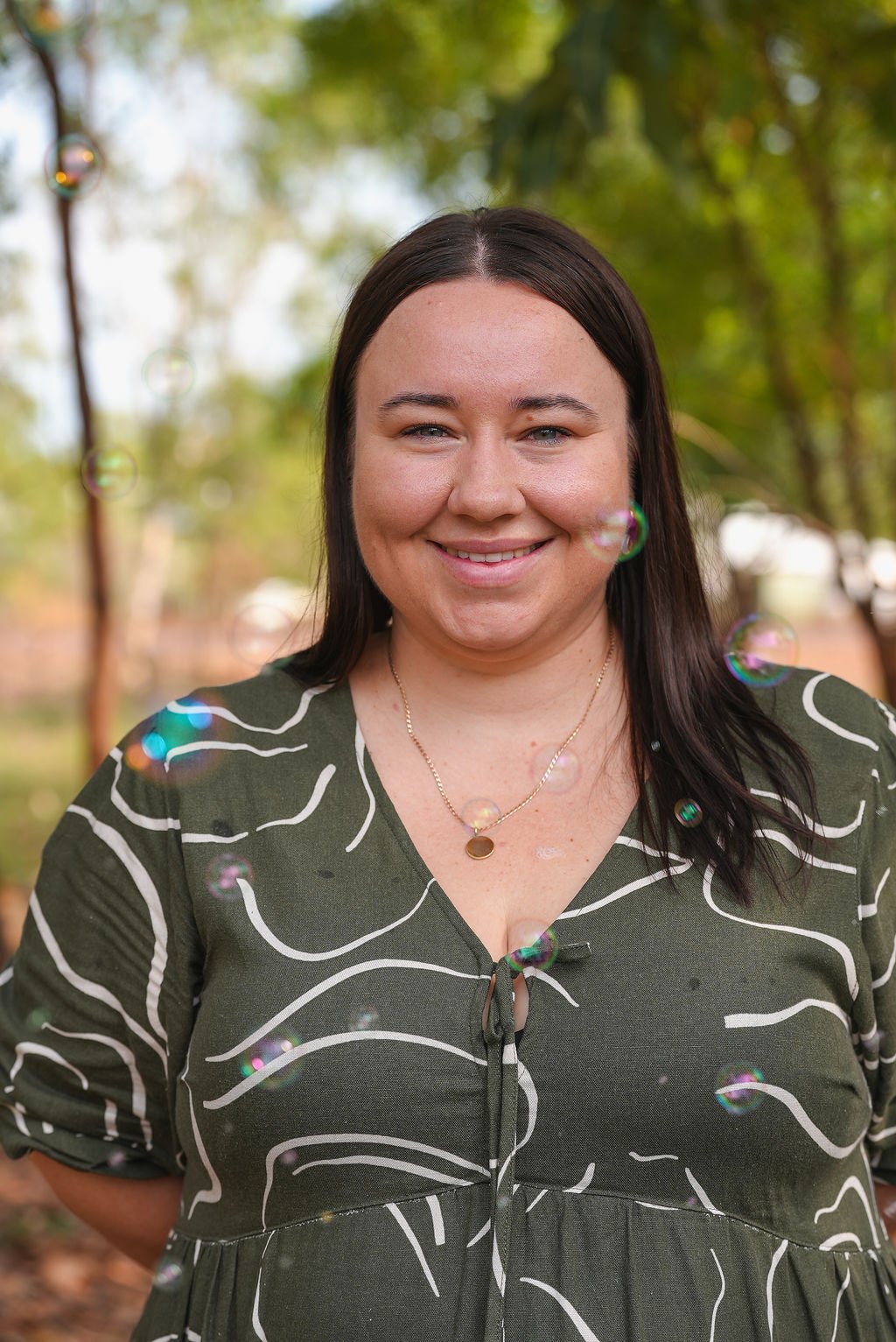 A woman with long dark hair blowing bubbles outdoors in a park with green trees in the background.