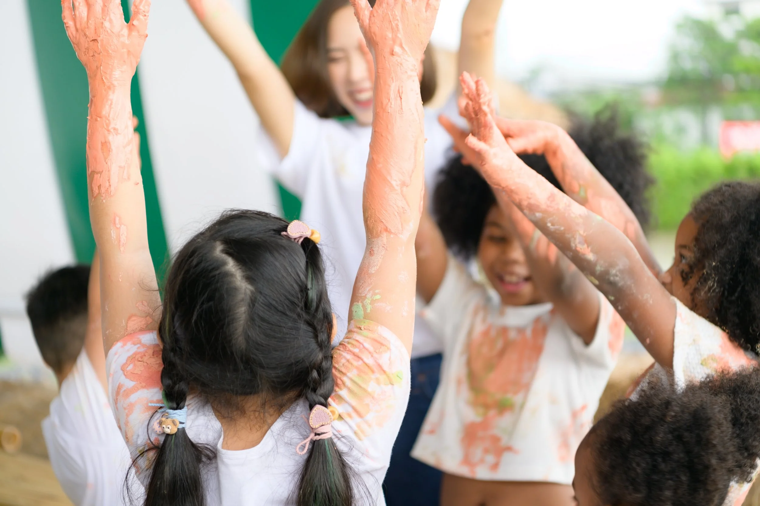 Children with painted arms celebrating and playing together outside, raising their arms in joy, with a supervisor smiling in the background.