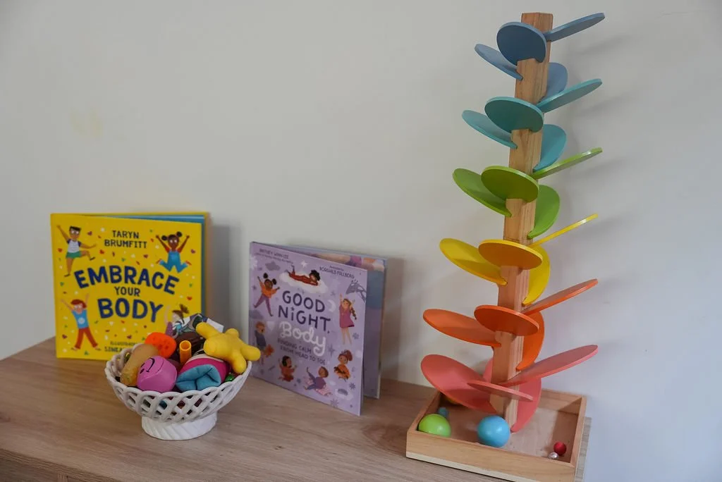 A wooden activity toy with colorful spinning discs, two books titled 'Embrace Your Body' and 'Good Night, Body,' and a white basket with toys on a wooden surface against a plain wall.